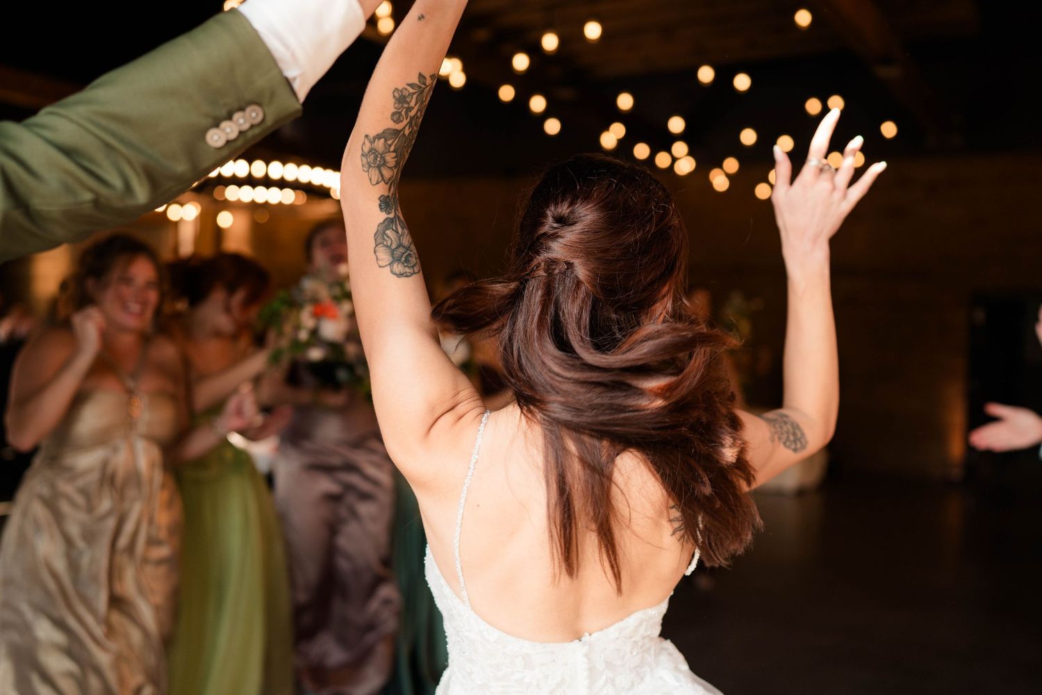 Bride with tattoos raises hands joyfully on wedding reception dance floor under warm string lights.