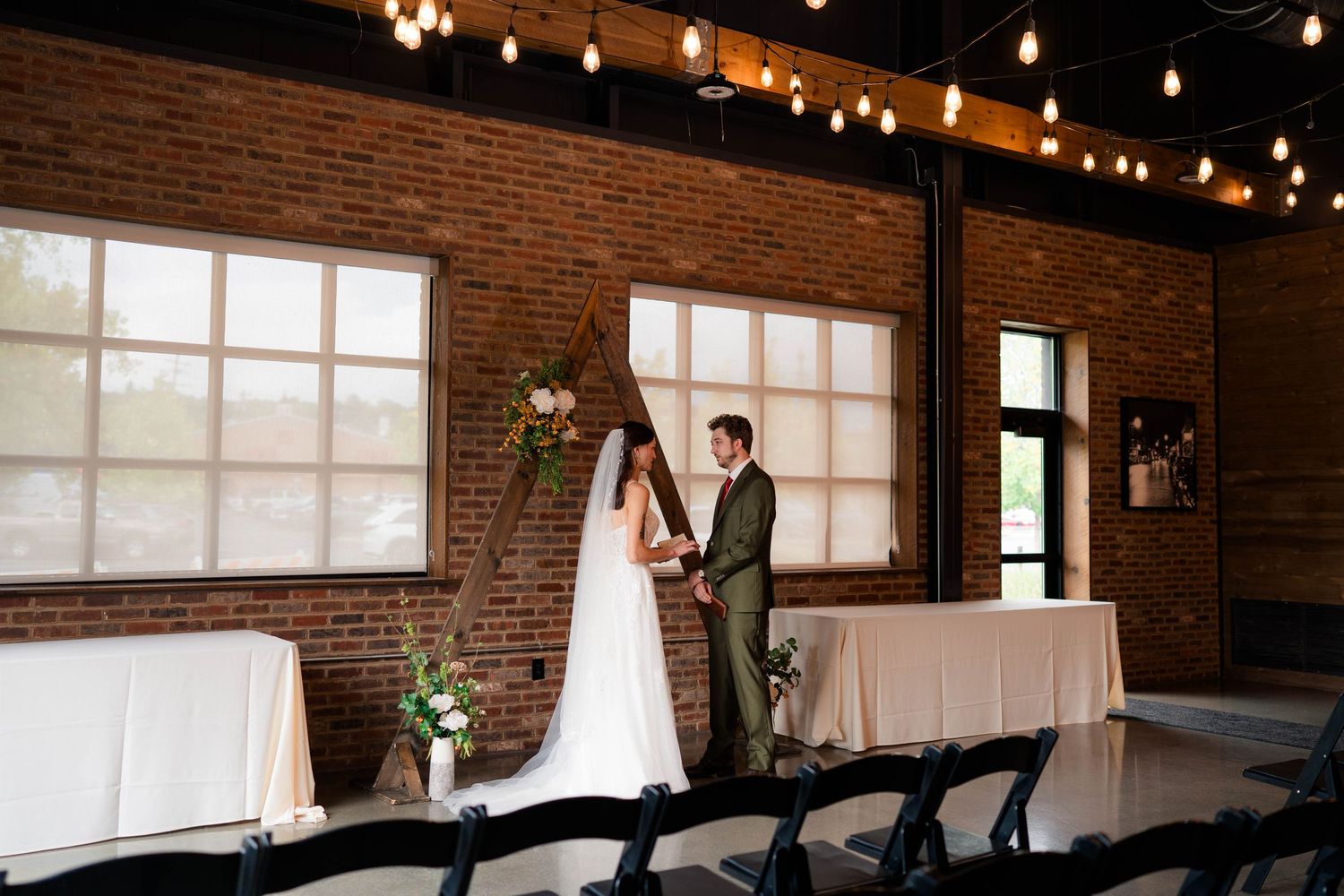 Bride and groom holding hands at decorated altar in industrial brick venue with string lights.