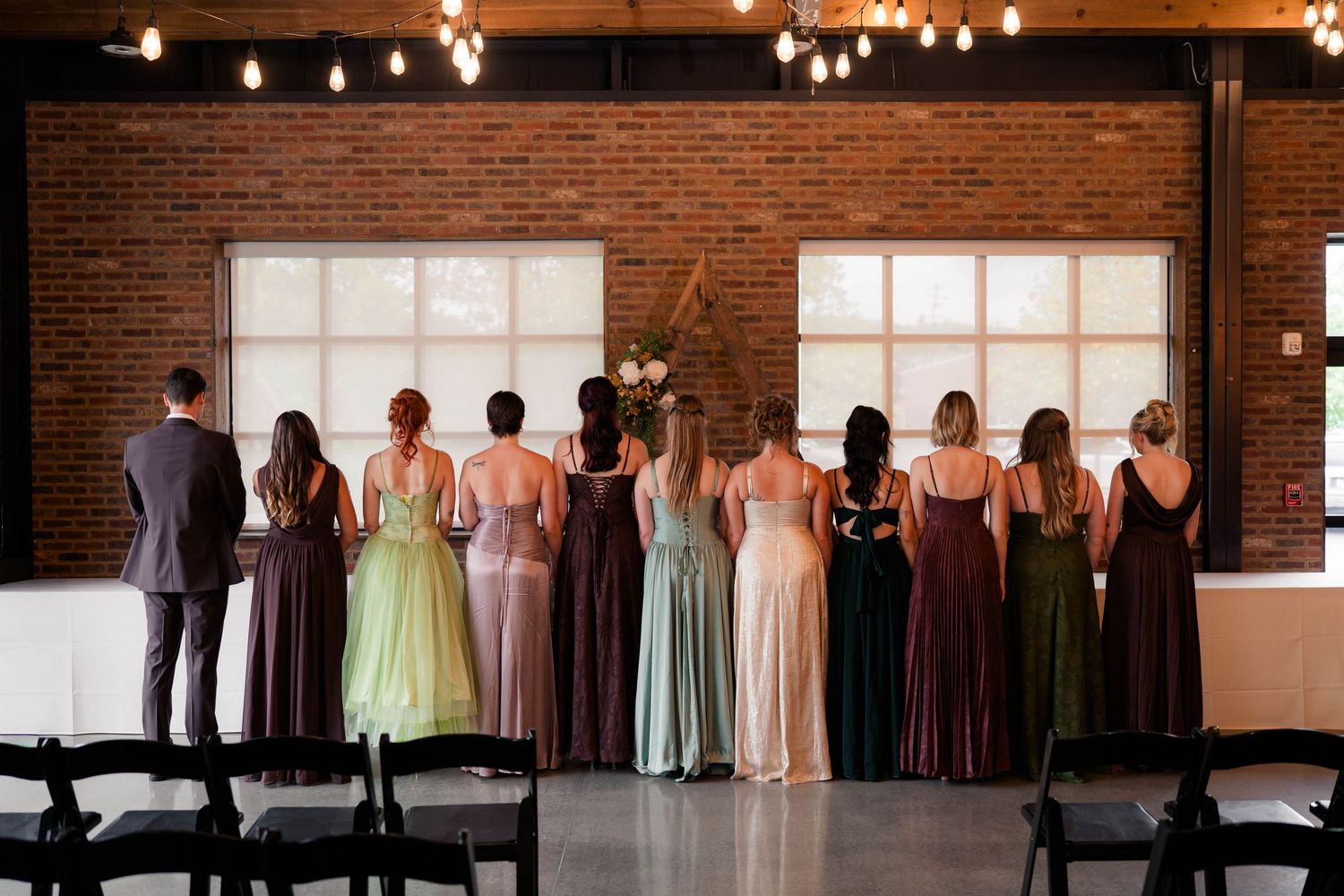 Wedding party standing in a row in colorful mismatched gowns inside a rustic brick venue with Edison bulb lighting.