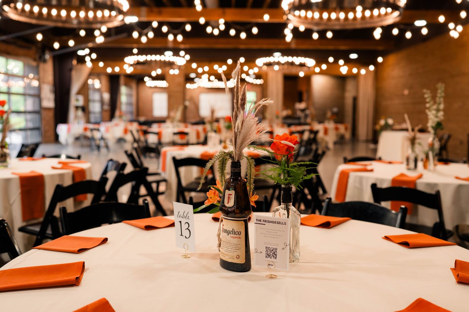 Elegant wedding reception venue with string lights, orange napkins, and wine bottle centerpieces.