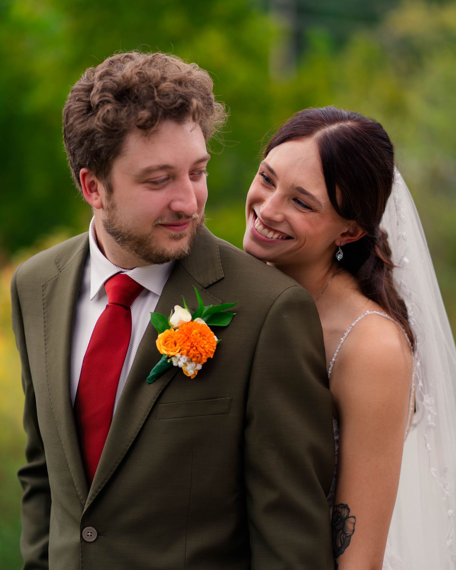 Bride in veil smiling at groom in olive green suit with orange boutonniere outdoors.