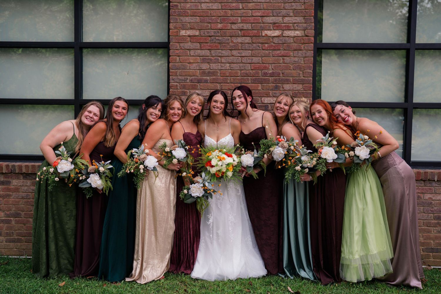 Bride surrounded by bridesmaids in mismatched earth-tone dresses holding colorful bouquets outdoors.