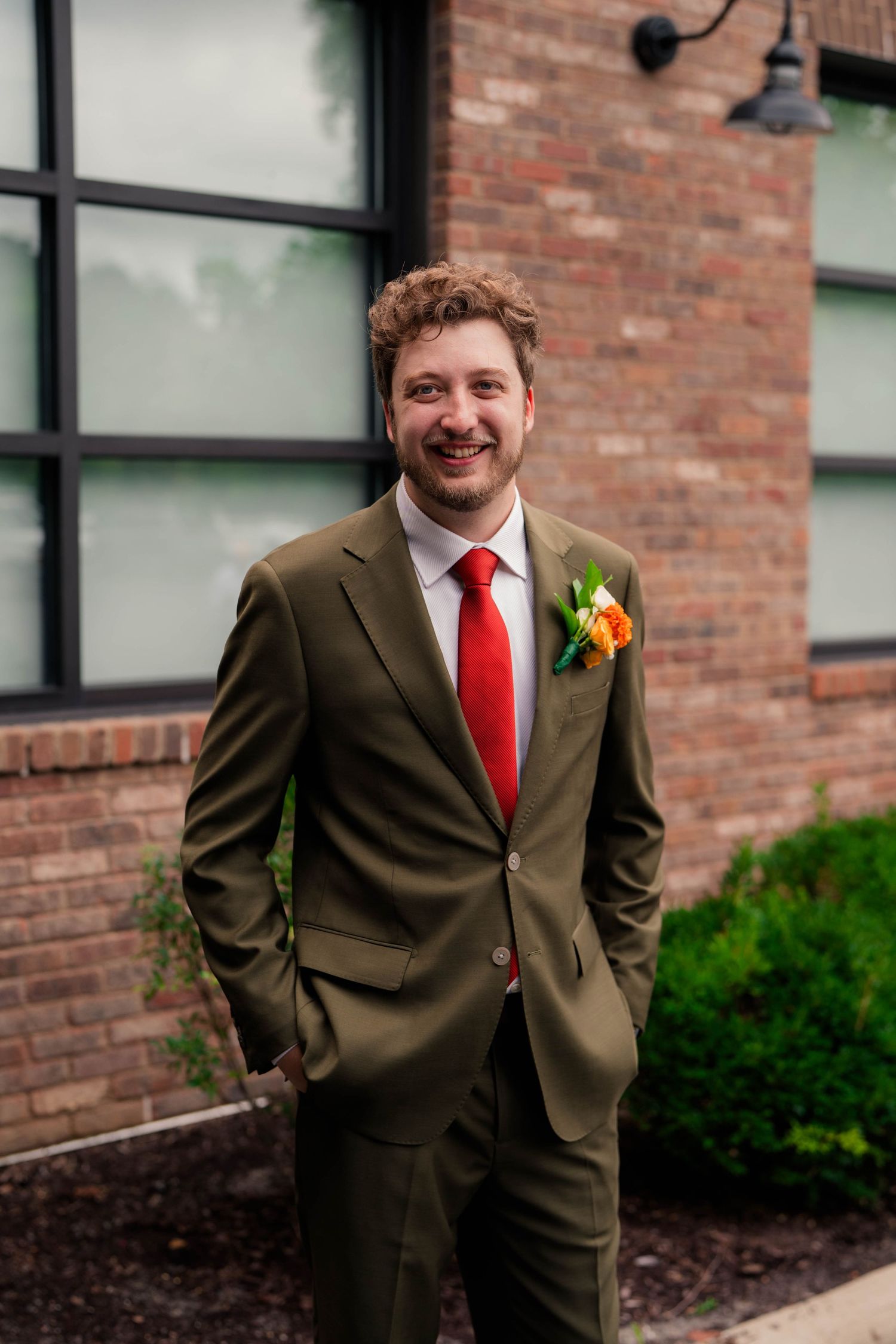 Smiling groom in olive green suit with red tie and orange boutonniere poses outside brick venue.
