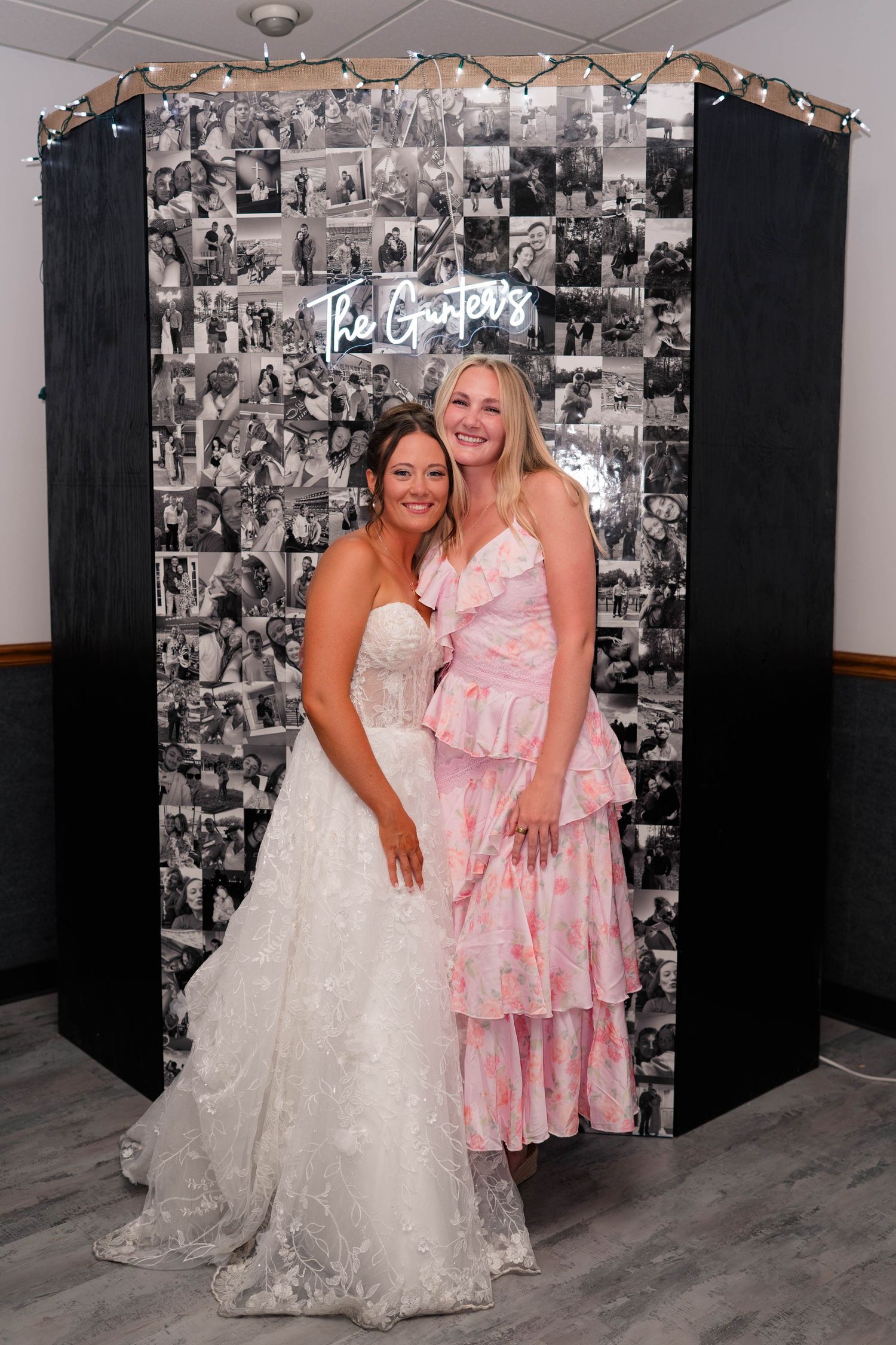 Bride in strapless white gown smiles with guest in pink ruffled dress before wedding photo collage backdrop.