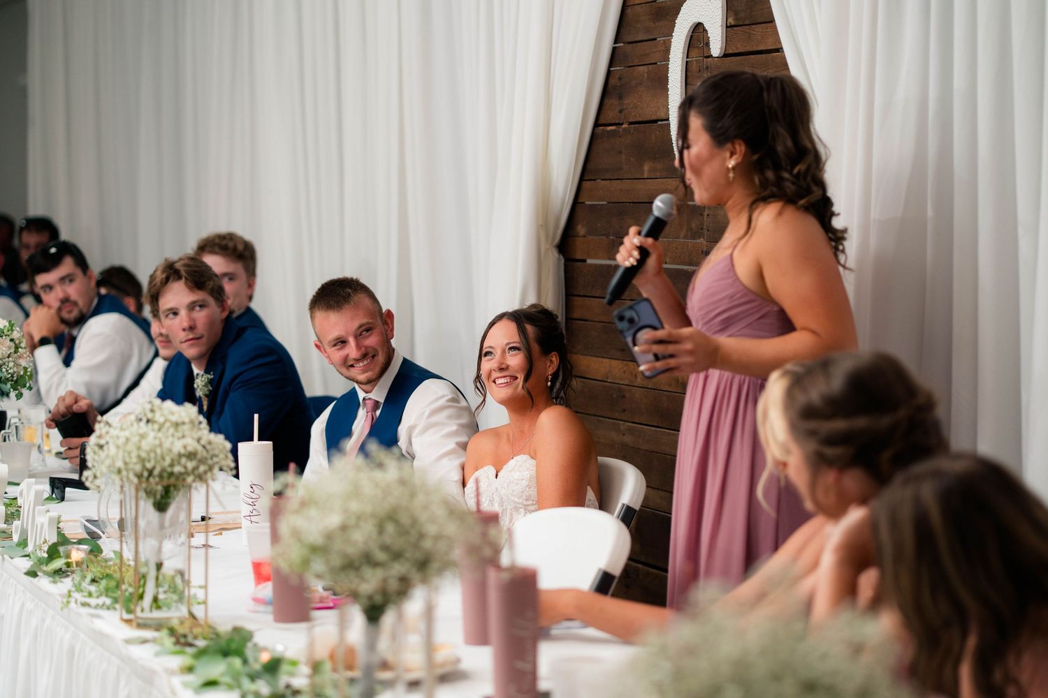 Bridesmaid in mauve dress delivers heartfelt speech to smiling bride and groom at wedding reception head table.