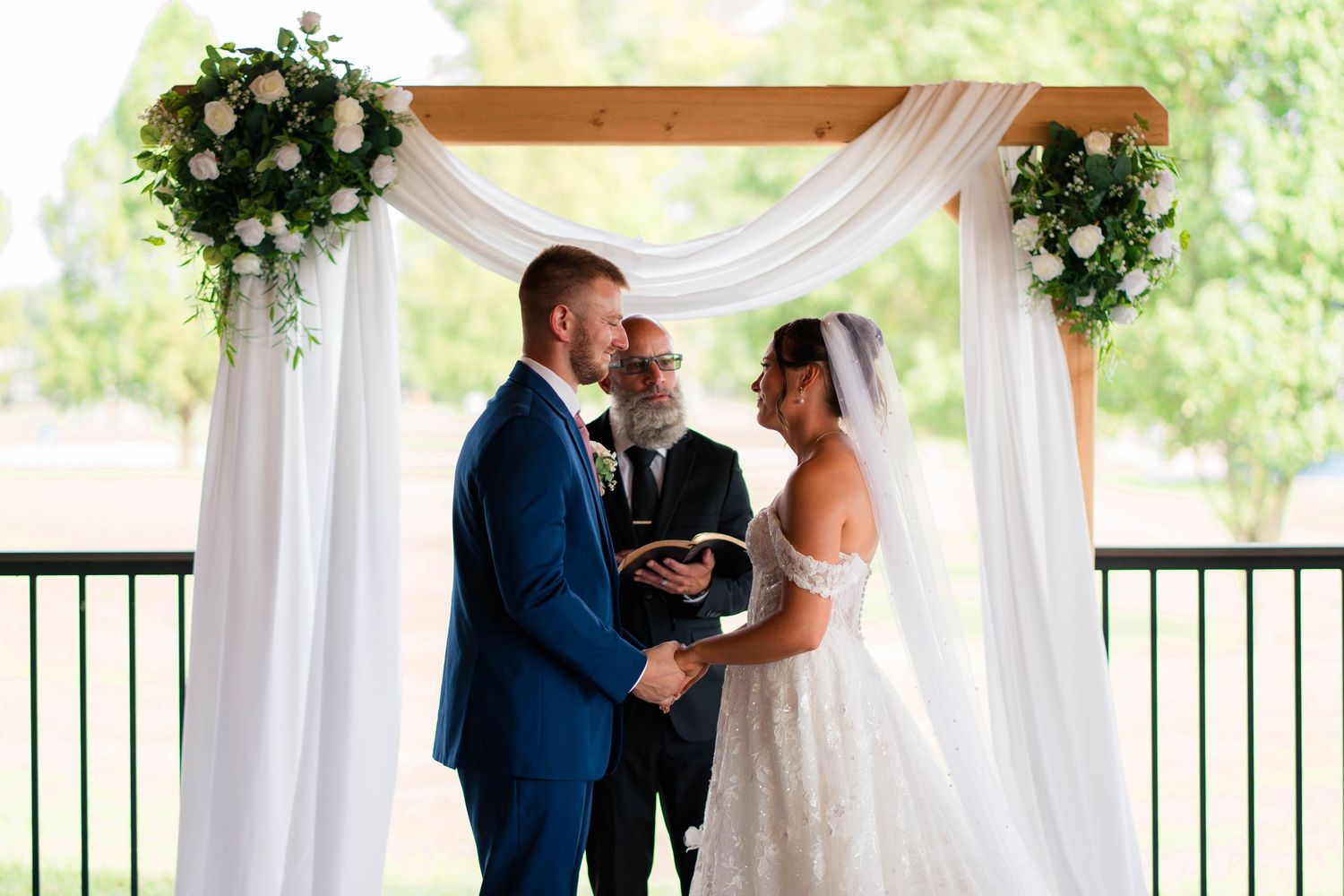Bride and groom exchange vows beneath a floral wooden arch at an elegant outdoor ceremony.