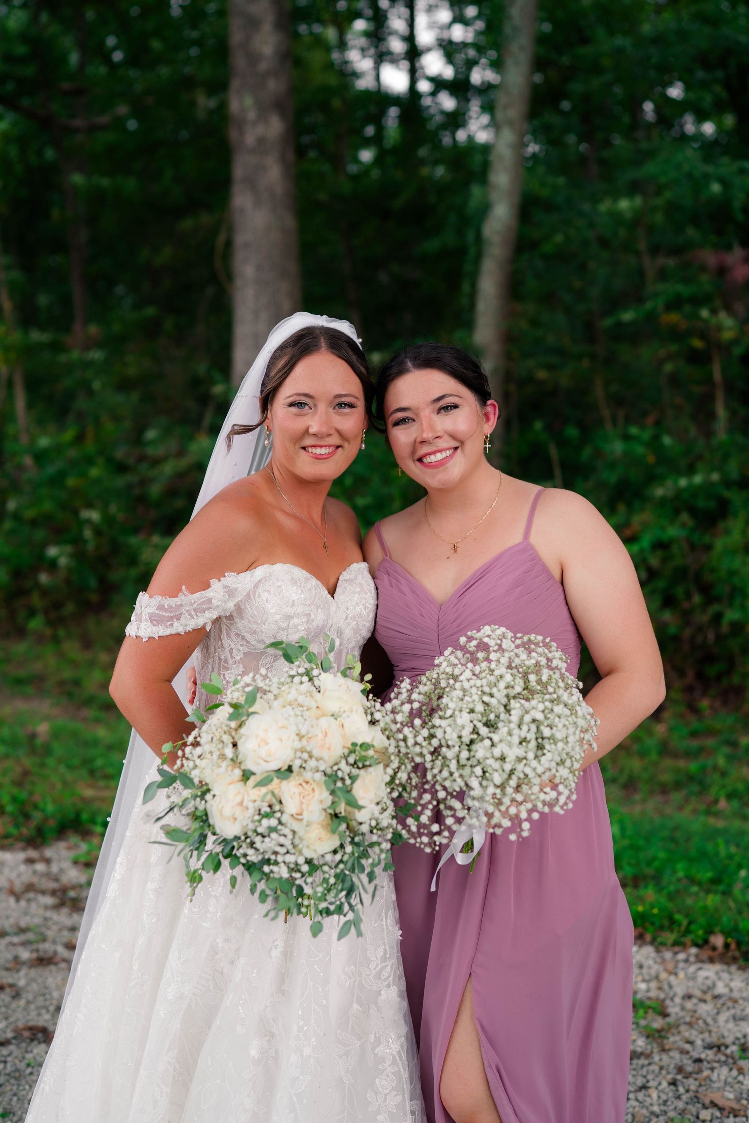 Smiling bride and bridesmaid in dusty rose gown holding white floral bouquets outdoors.