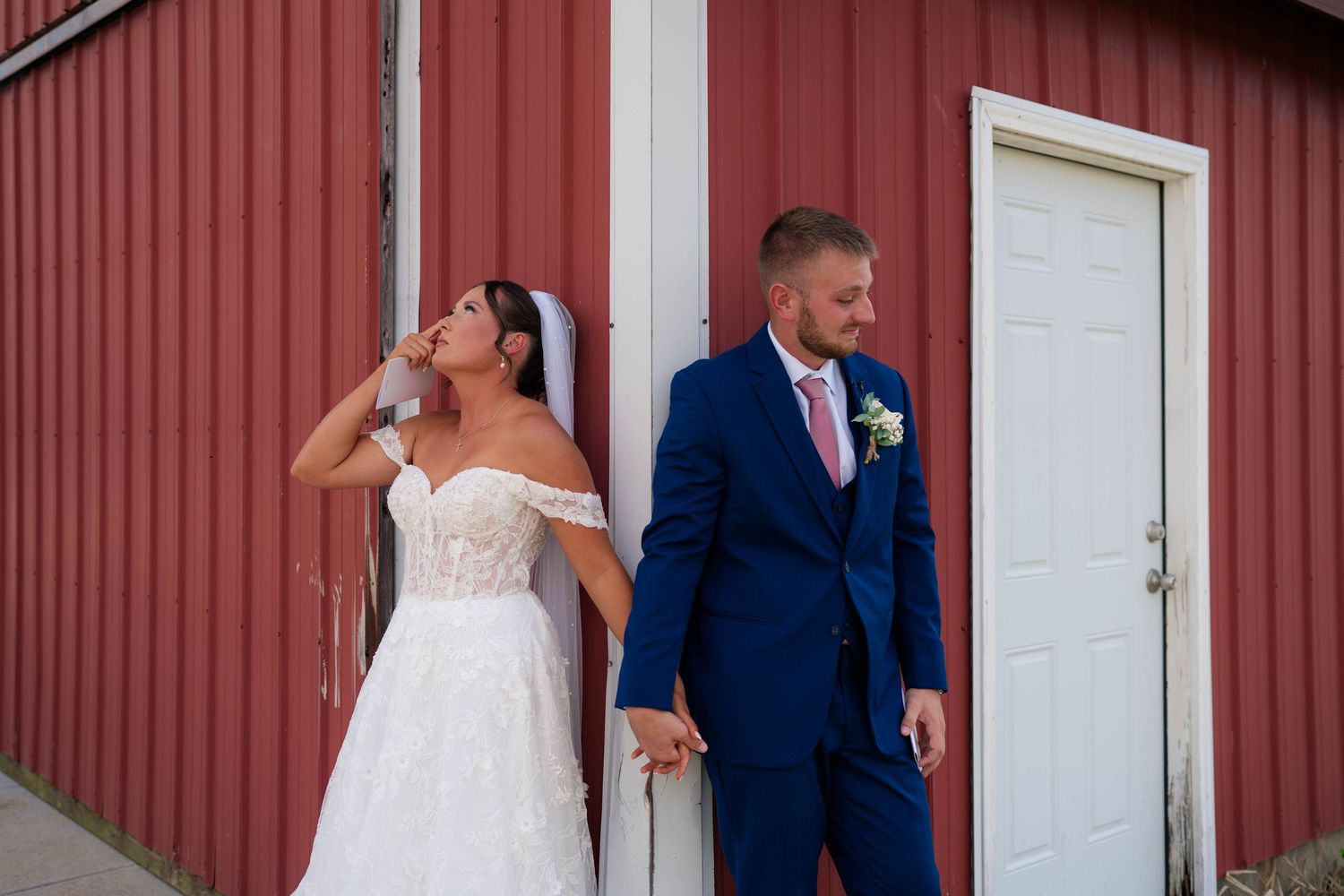 A bride and groom hold hands leaning against a red barn wall, she sips a drink as he looks away.