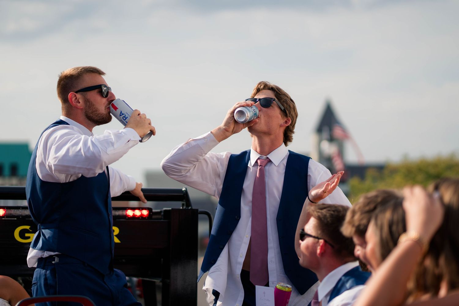 Two groomsmen in navy vests drinking canned beverages outdoors at a sunny wedding reception.