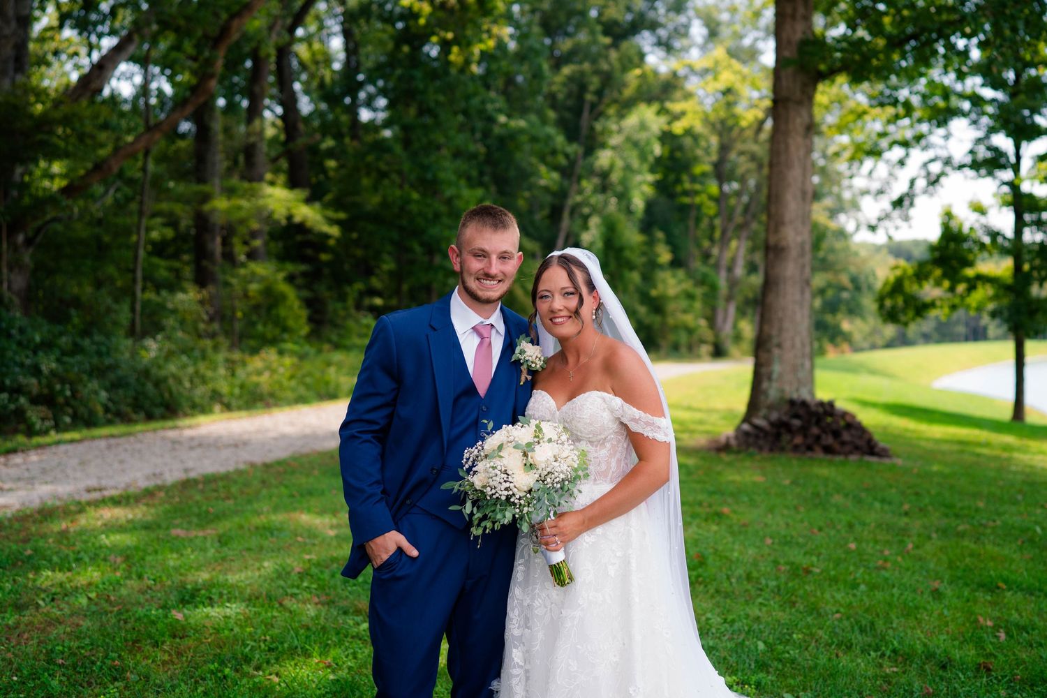 Smiling bride and groom pose together outdoors, groom in navy suit, bride holding white bouquet.