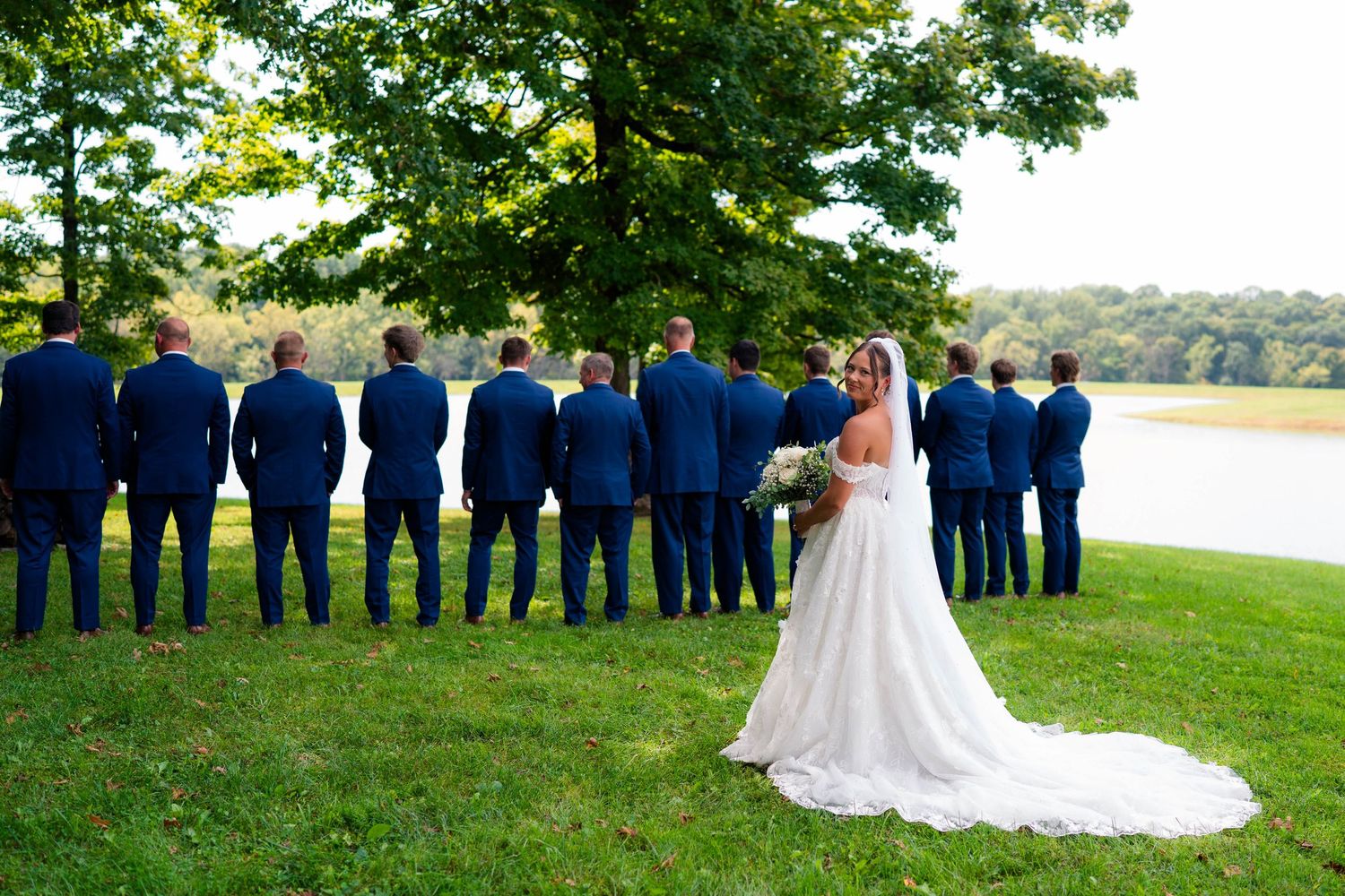 Bride in lace gown stands with groomsmen in navy suits facing scenic lakeside backdrop.