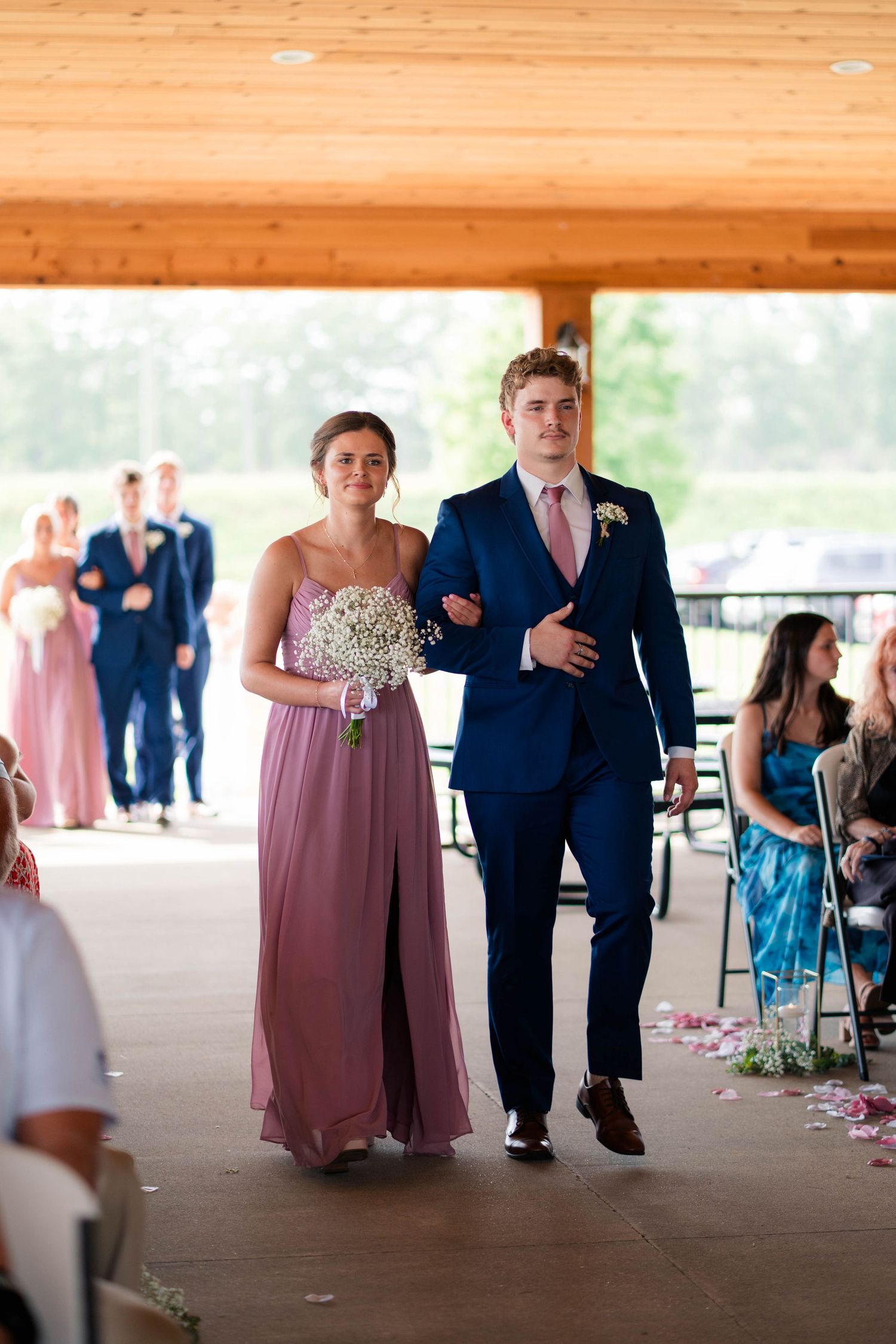 Bridesmaid in mauve gown and groomsman in navy suit walking down wedding aisle.
