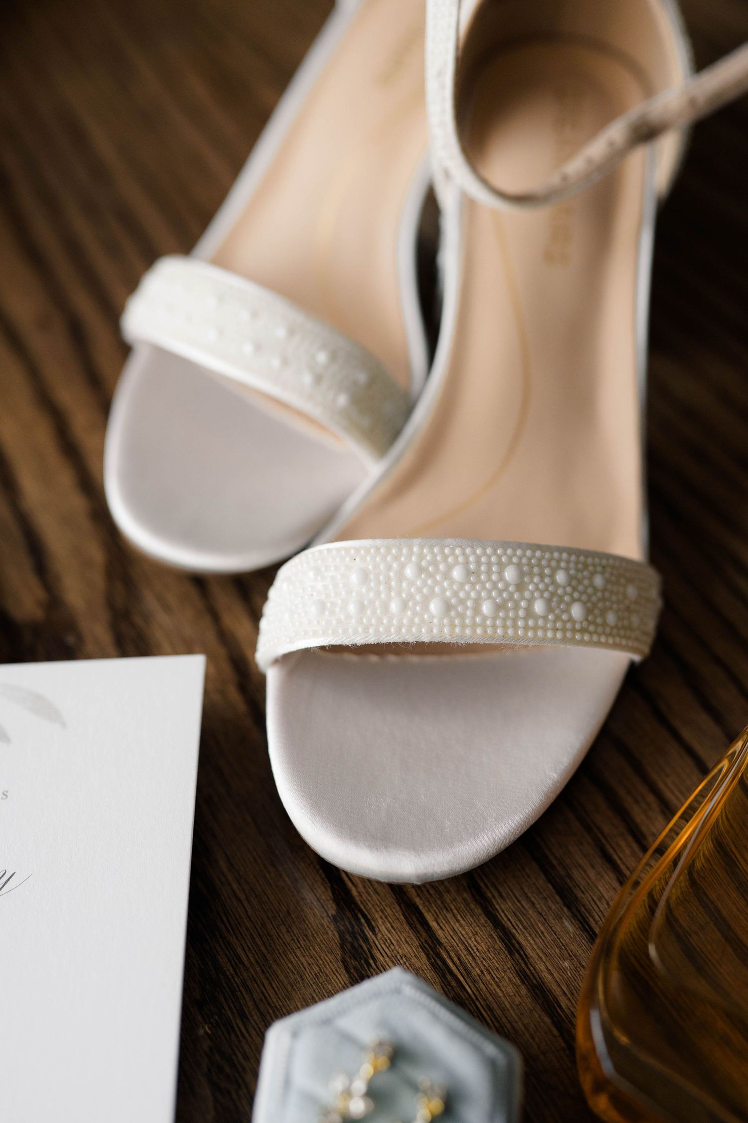 White pearl-embellished bridal heels on wooden surface beside wedding stationery and perfume bottle.
