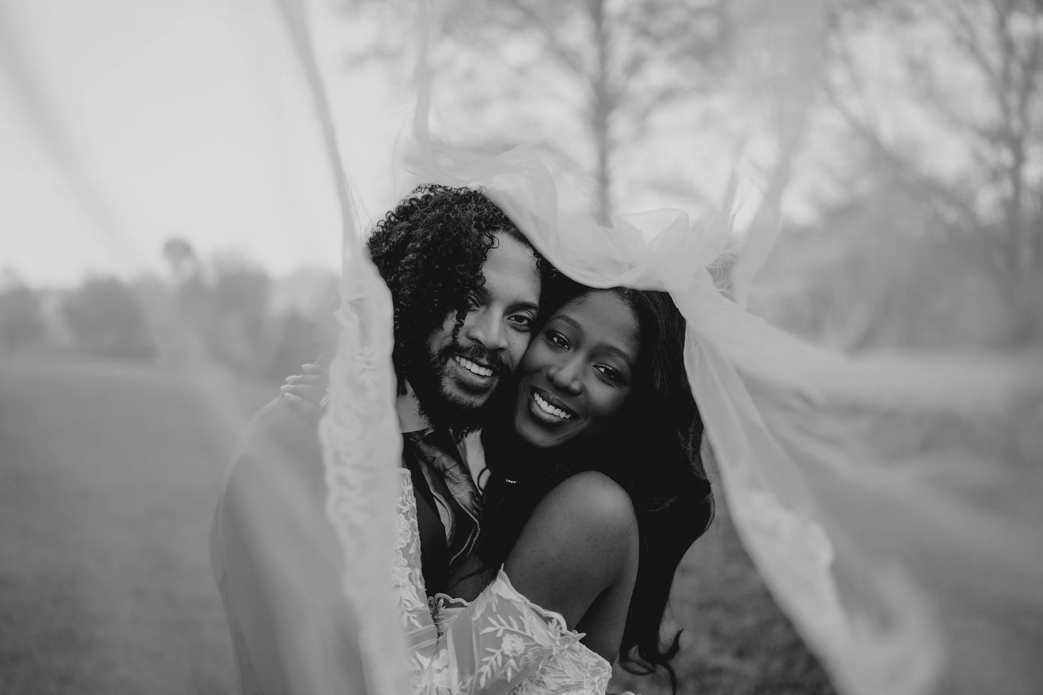 Black and white photo of a smiling bride and groom embracing under a flowing wedding veil outdoors.