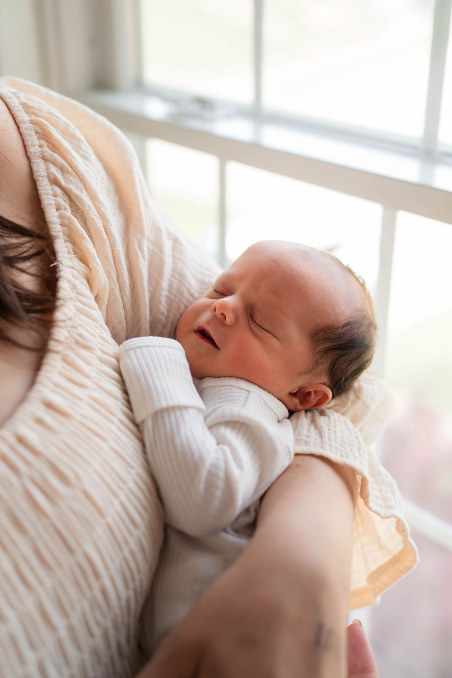 Peaceful newborn baby sleeping in mothers arms, wrapped in a soft white onesie near a bright window.