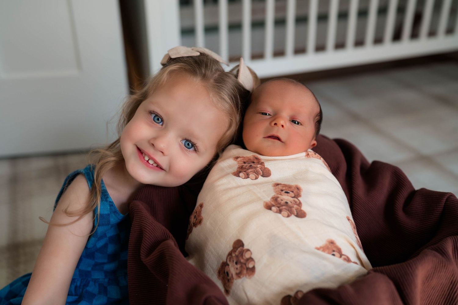Smiling blue-eyed toddler girl posing beside swaddled newborn baby wrapped in a teddy bear blanket.