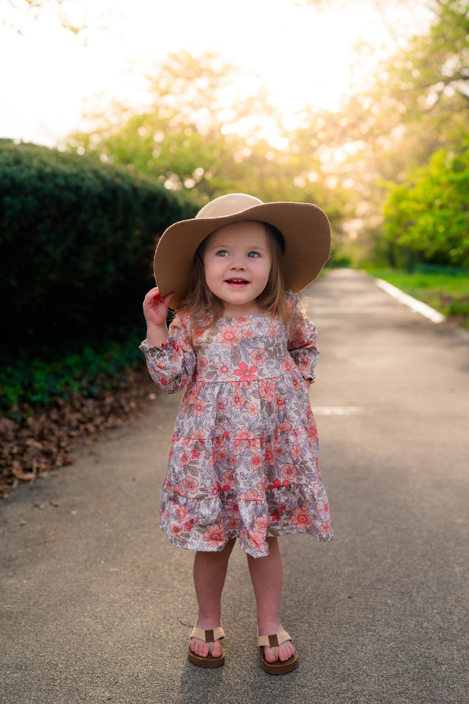 An adorable toddler girl in a floral dress and oversized sun hat smiles on a sunlit garden pathway.
