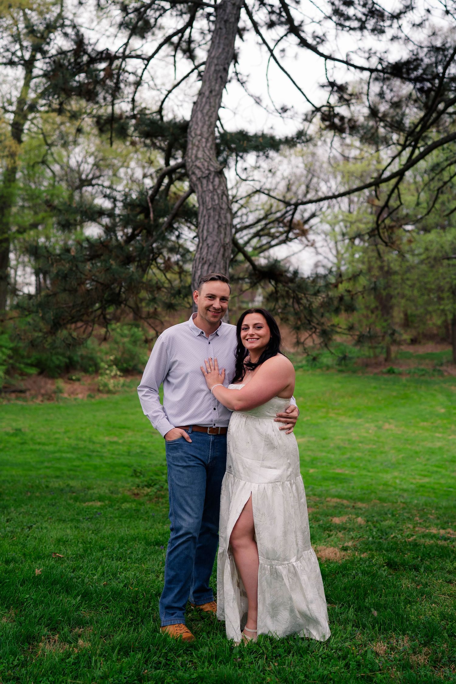 A happy couple embraces and smiles together on a lush green lawn beneath a large pine tree.
