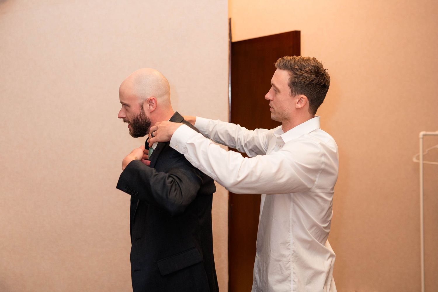 Man in white shirt adjusting the collar of a bald man in a dark suit jacket indoors.