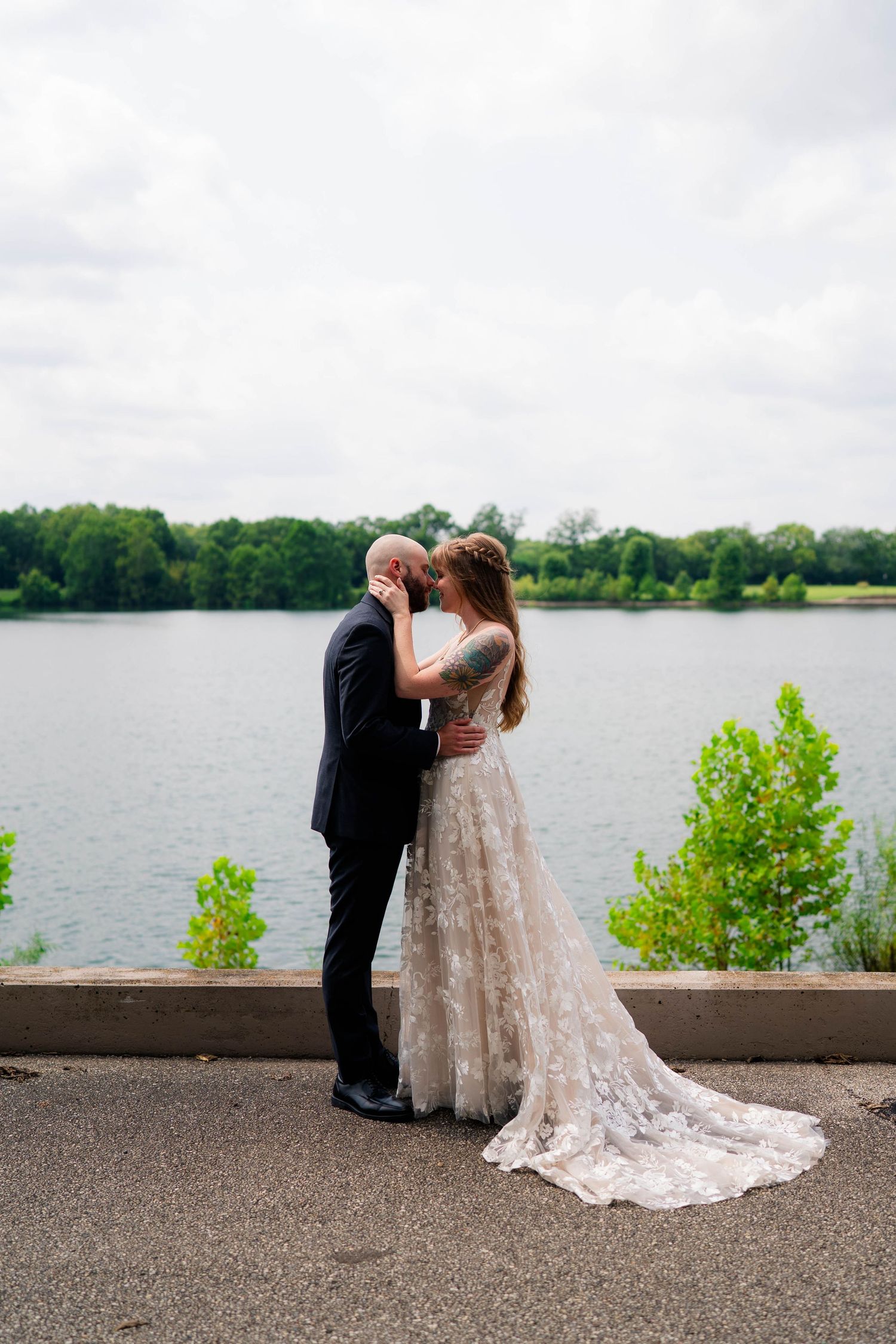 Bride and groom share a romantic kiss by a serene lake, bride wearing a lace gown with a long train.