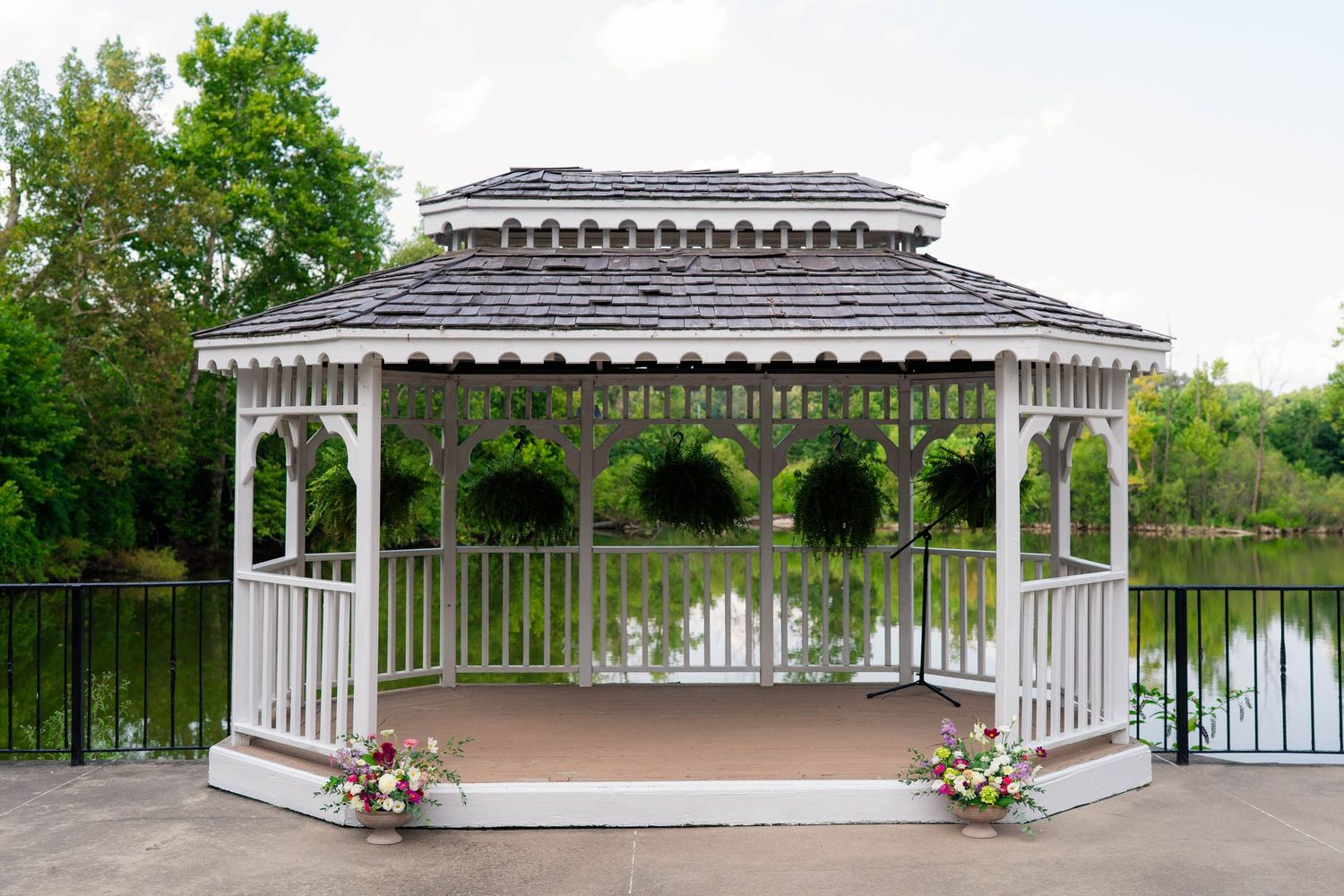 Elegant white gazebo wedding ceremony venue overlooking a serene pond, decorated with floral arrangements and hanging plants.