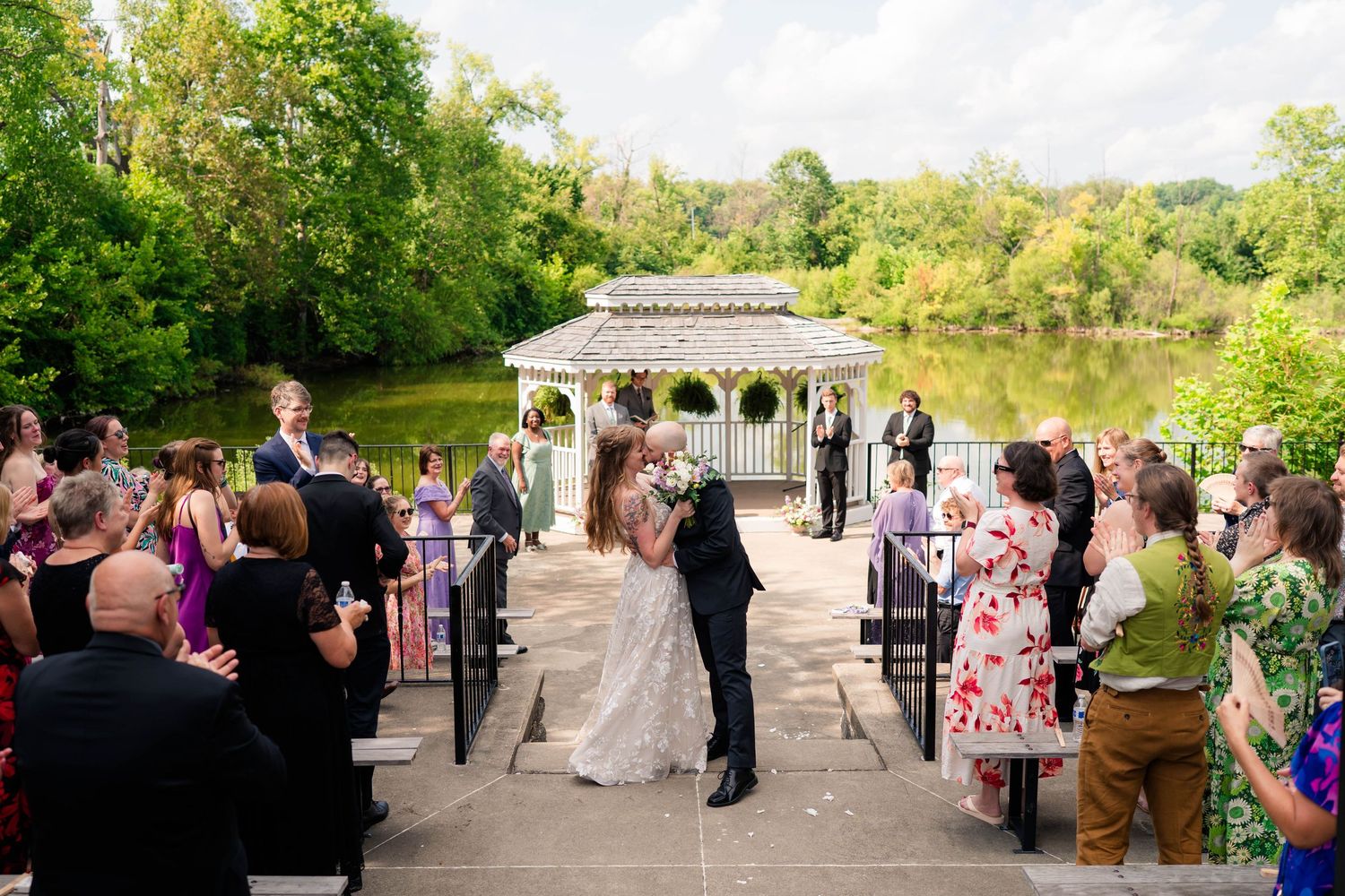 Bride and groom share first kiss at outdoor wedding ceremony by a scenic lakeside gazebo.