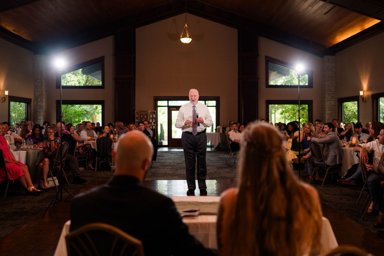 Man giving a toast speech at a large indoor wedding reception venue.