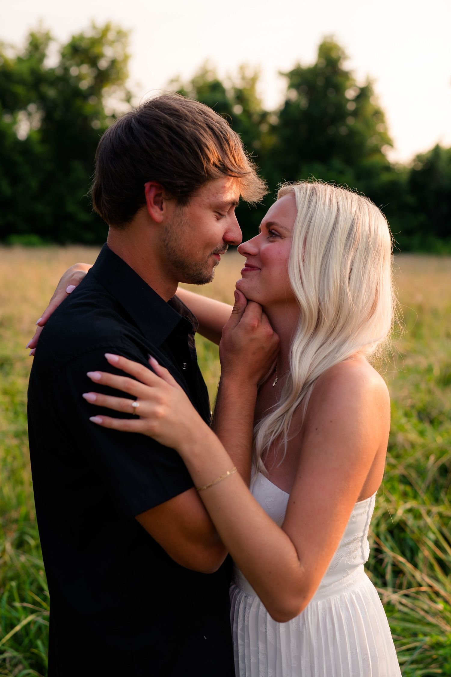 A couple shares a romantic moment in a golden field, the blonde woman in white tenderly holding her partner's face close to hers.