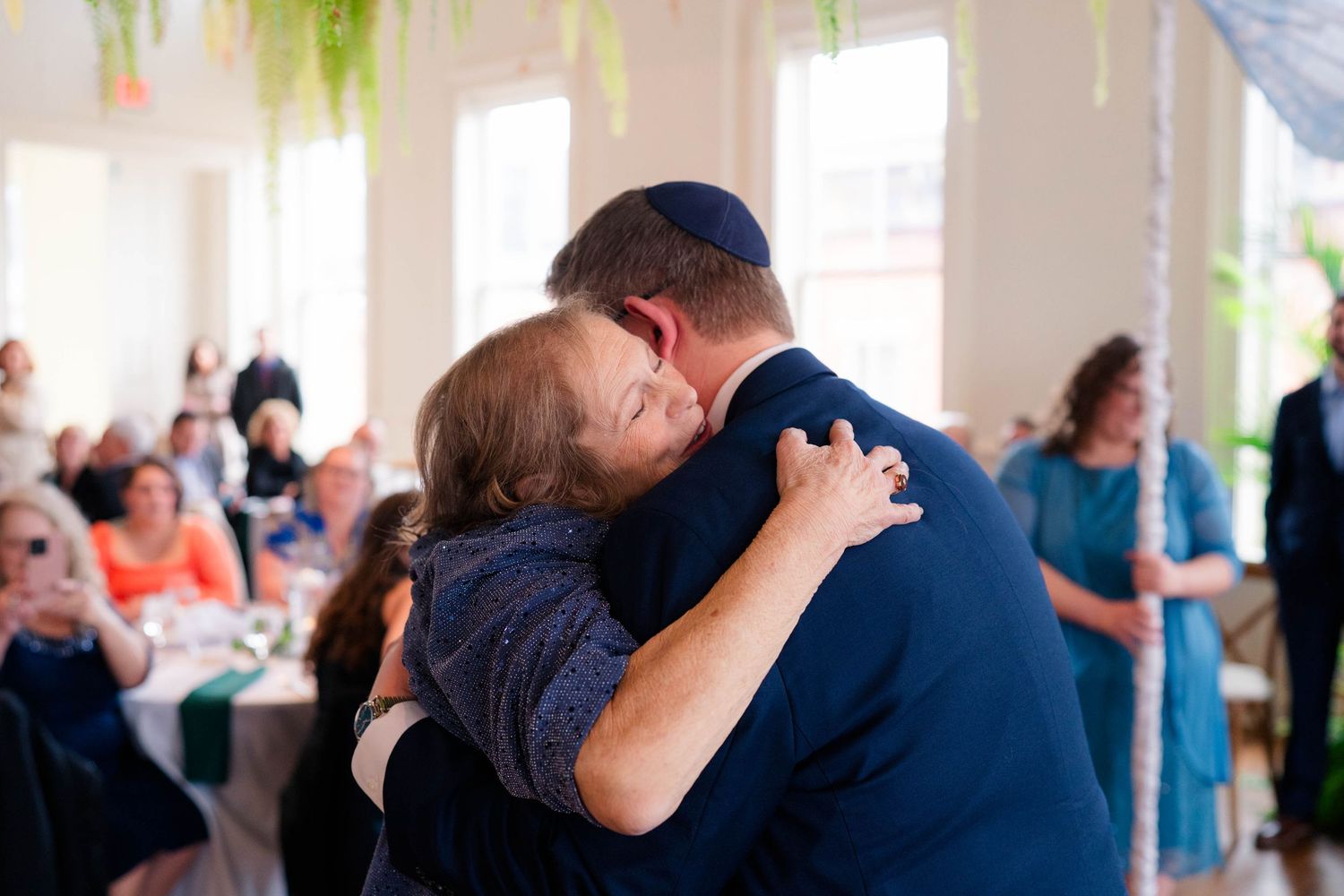 Groom in navy suit and kippah shares emotional hug with mother during wedding reception dance.