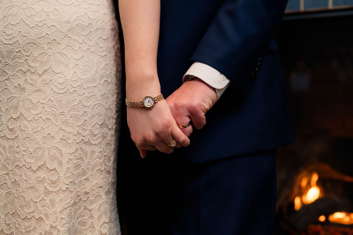 Close-up of bride and groom holding hands near a fireplace, bride wearing a lace dress and gold watch.