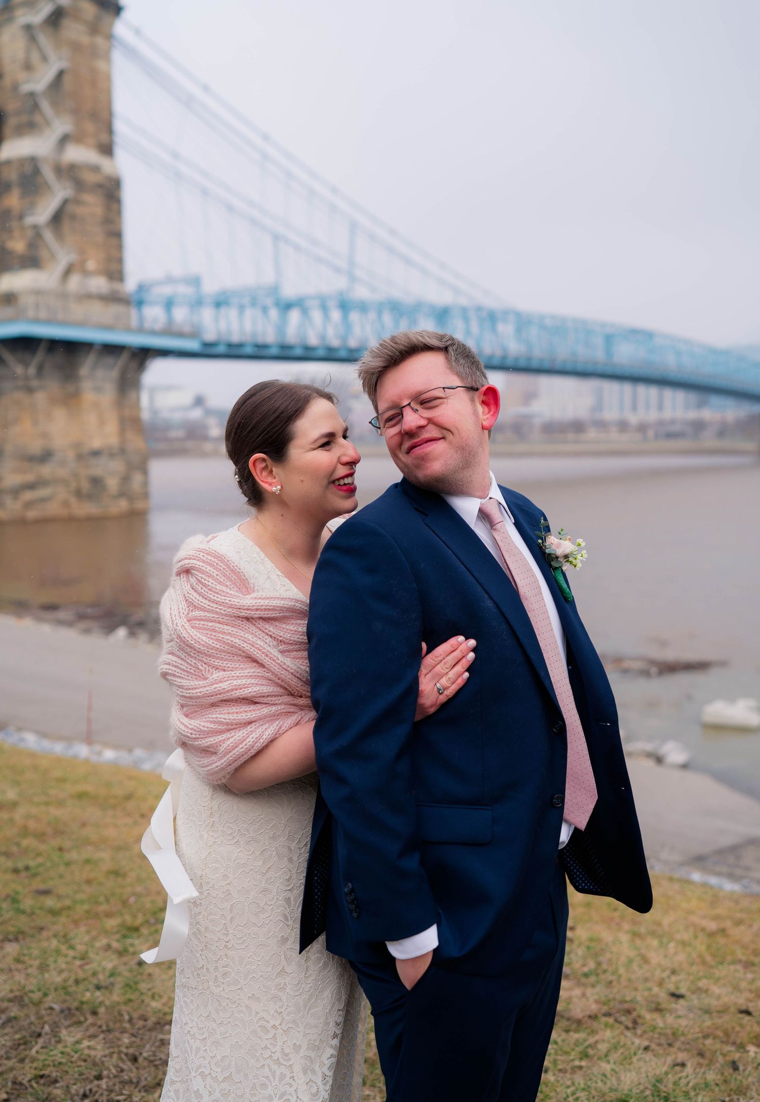 Bride and groom smiling together near a blue suspension bridge over a river on their winter wedding day.