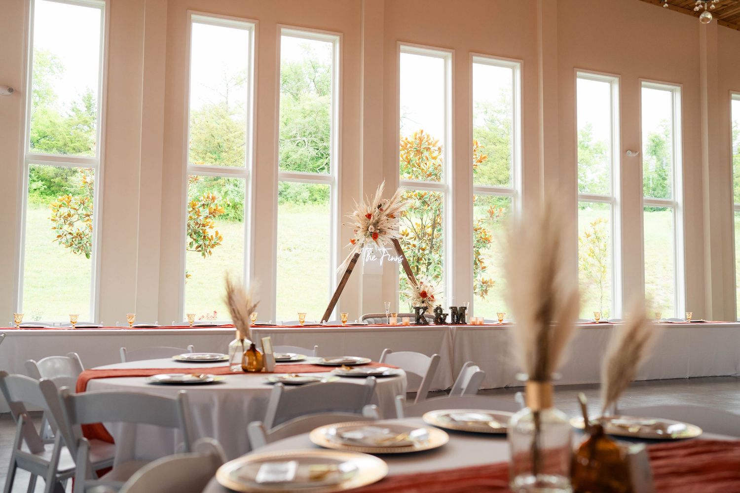 Elegant wedding reception hall with tall windows, round tables, and pampas grass centerpieces.