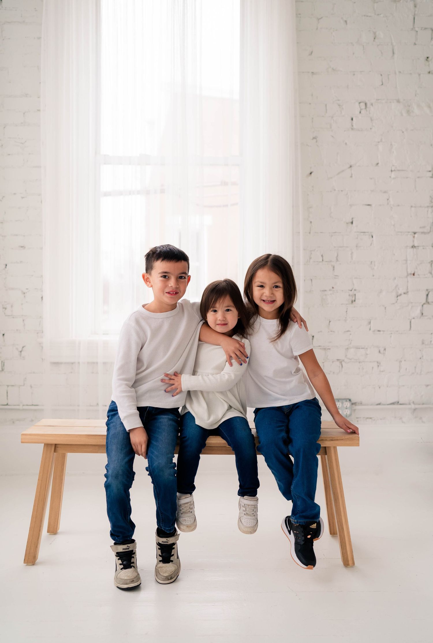 Three children in white tops and jeans sitting together on a wooden bench in a bright studio.
