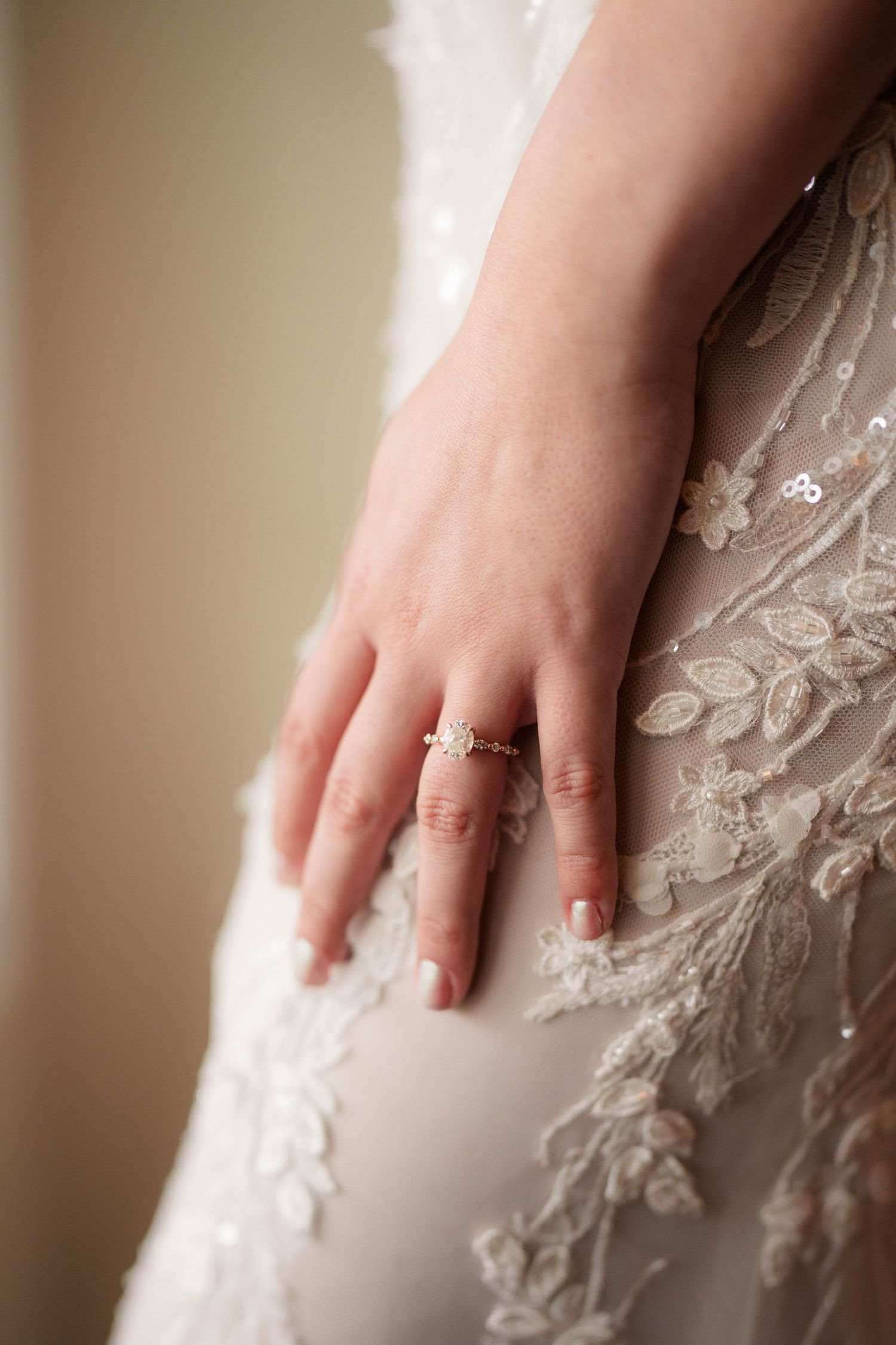 Close-up of a bride's hand with a delicate diamond ring resting on her intricate lace wedding gown.