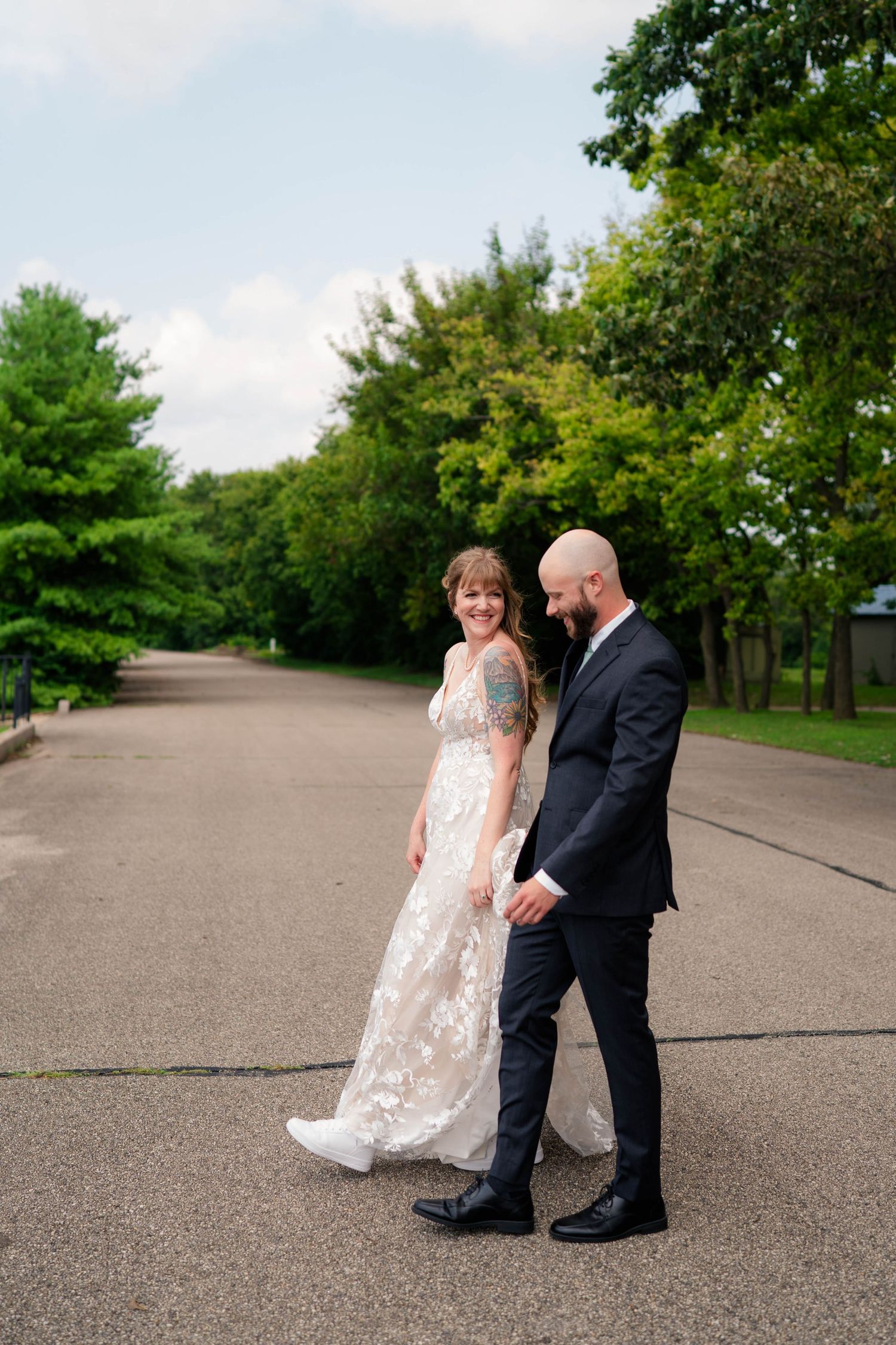 Smiling bride in lace gown and tattooed groom in dark suit walking together on an outdoor road surrounded by trees.