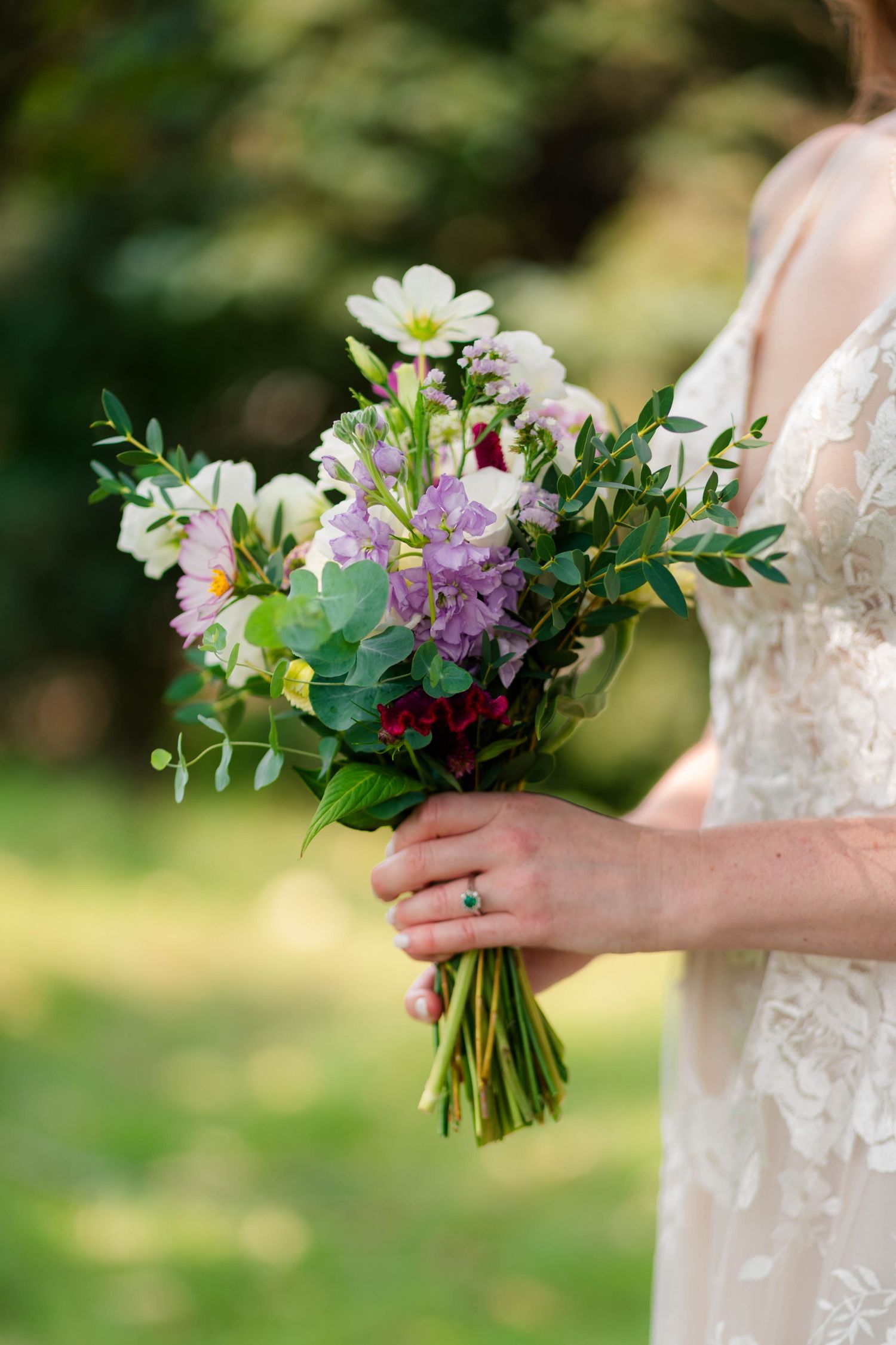 A bride holds a wildflower bouquet with white, purple, and red blooms with greenery outdoors.