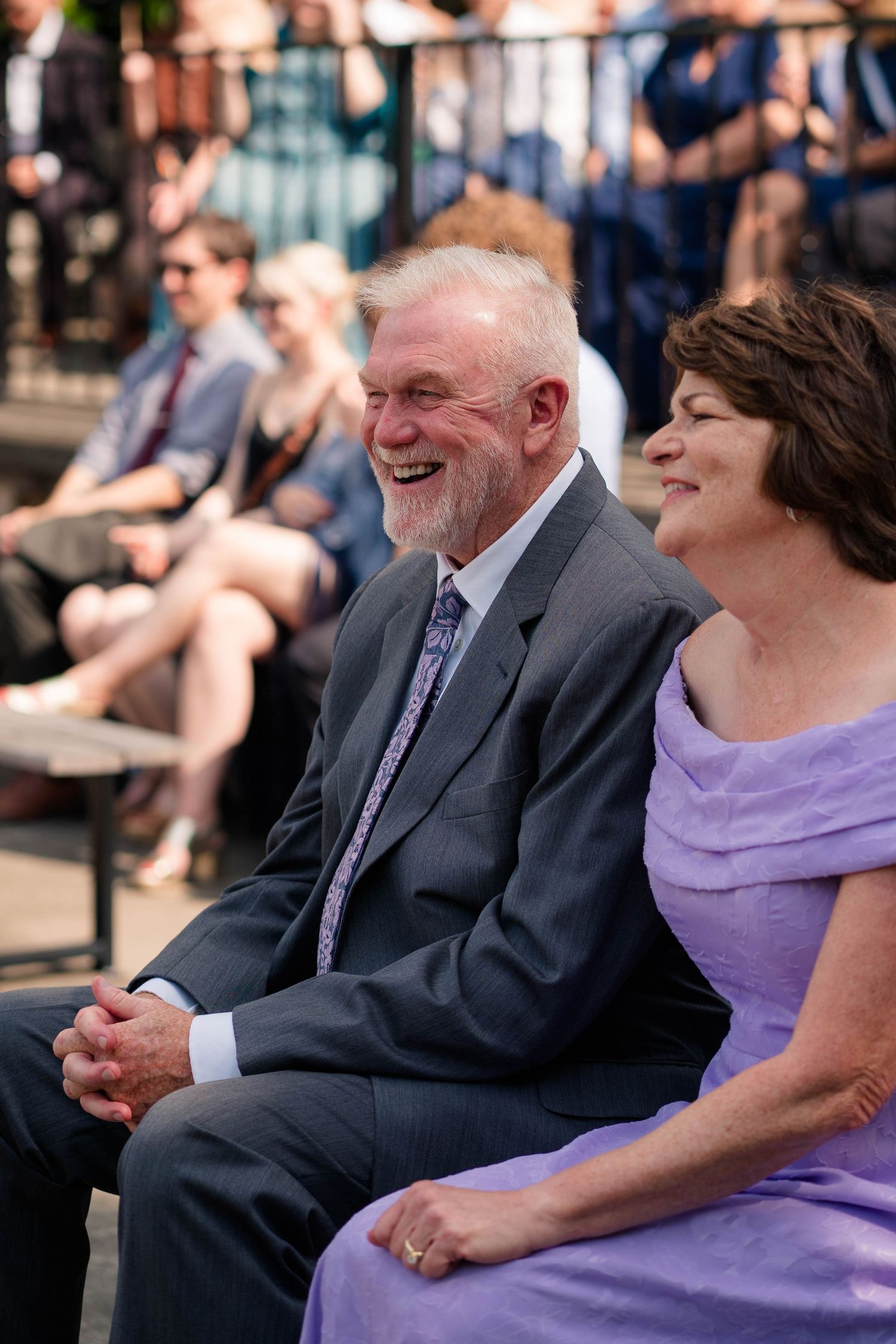 An elderly couple in formal attire laughs joyfully while seated at an outdoor wedding ceremony.