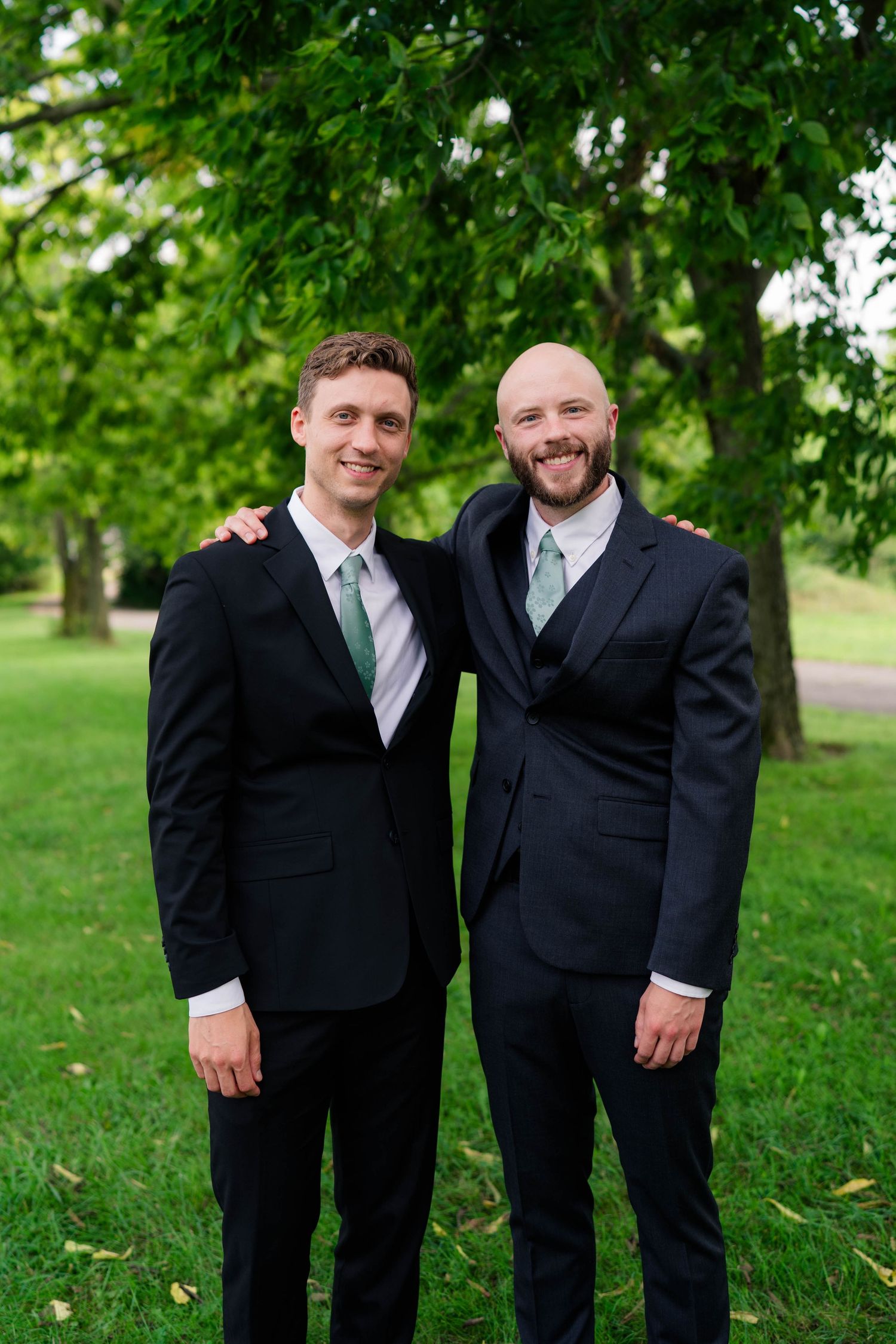 Two men in black suits with green ties smiling together at outdoor wedding celebration.