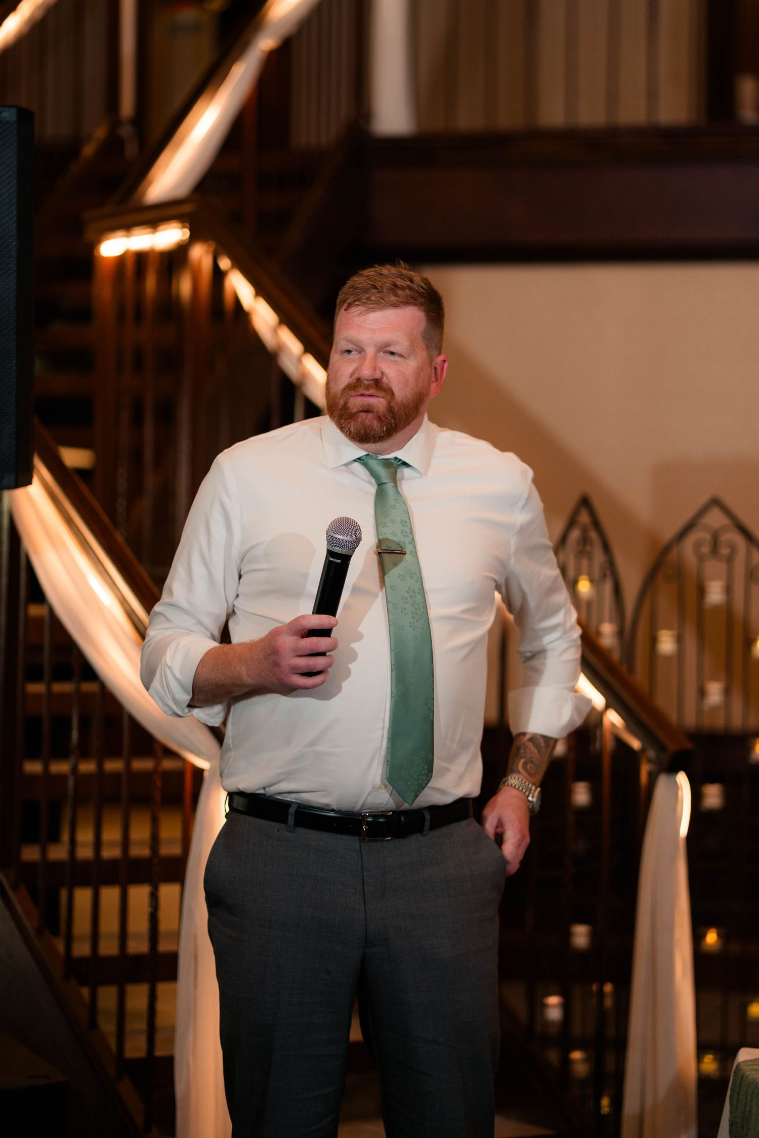 Red-bearded man in white shirt and green tie holds microphone giving a speech at wedding reception.