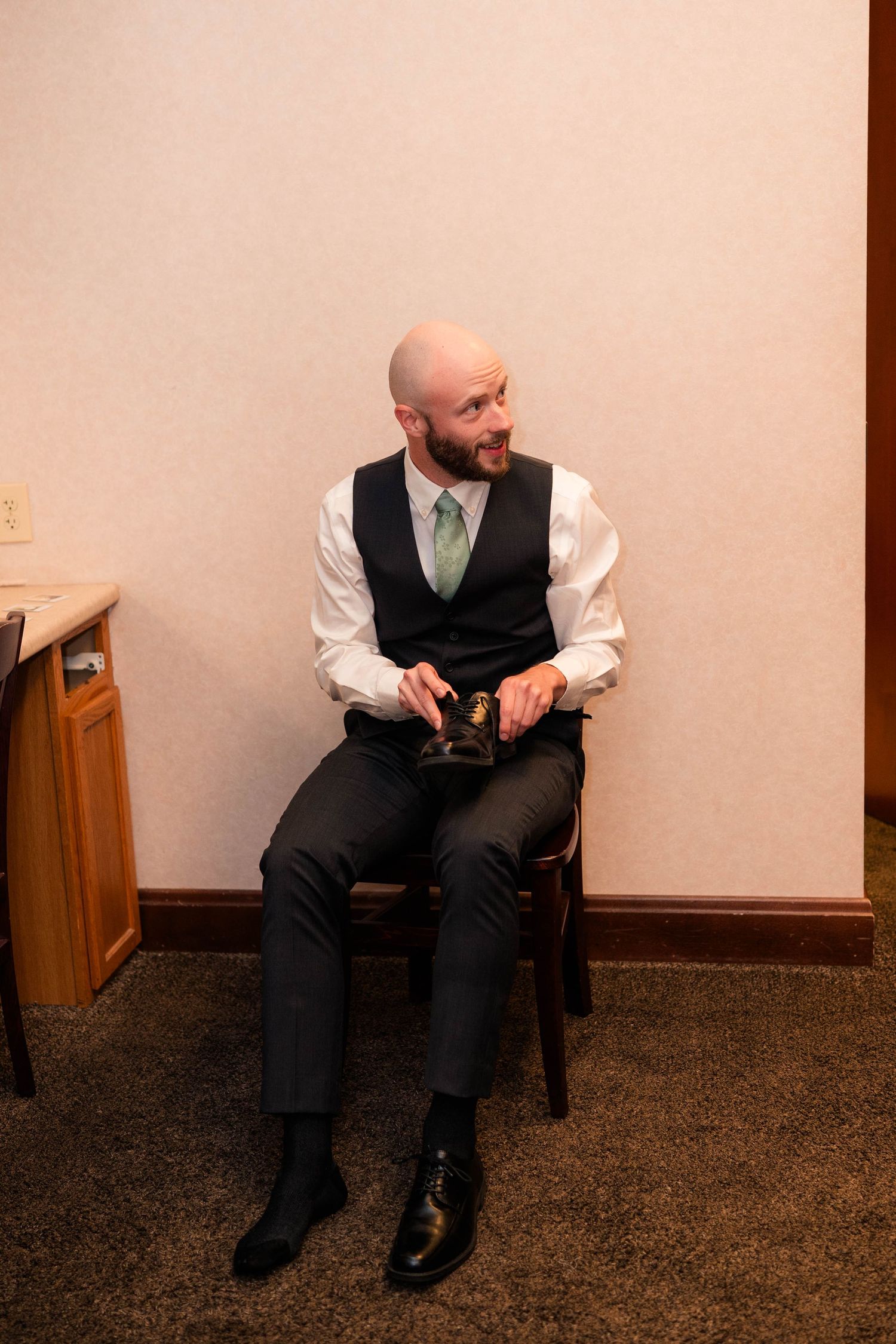 Bald groom in black vest and green tie sits on chair holding shoes while getting ready for wedding.
