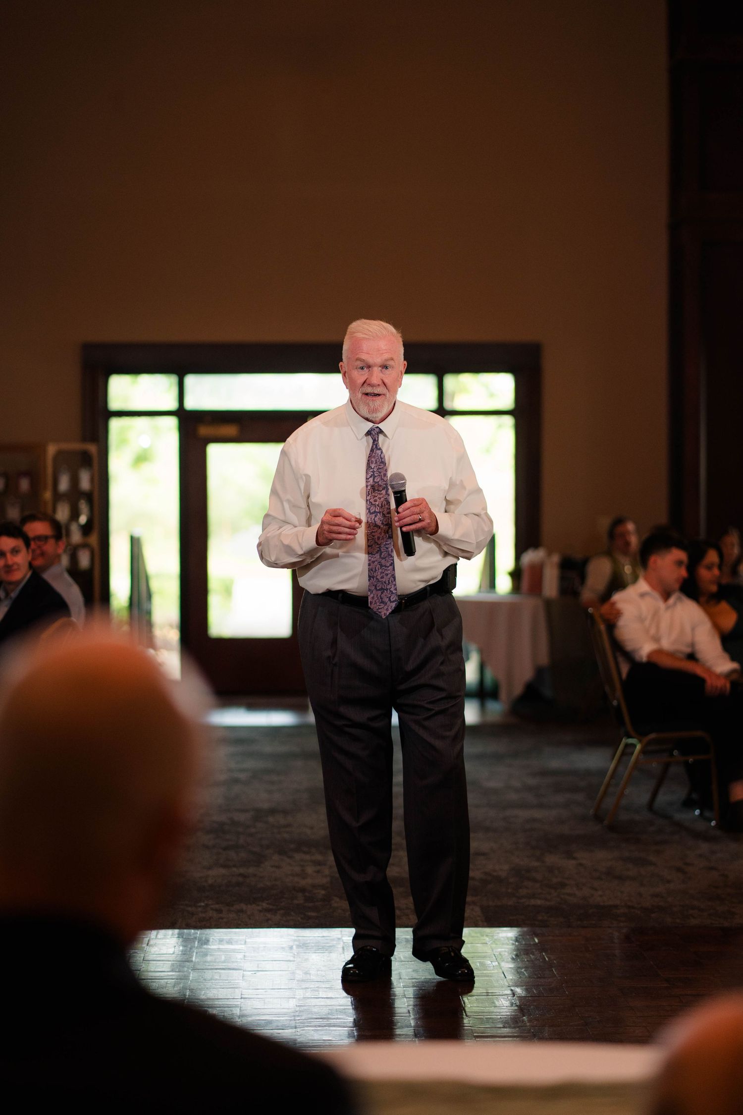 An older man in a white dress shirt and tie speaks into a microphone during a wedding reception speech.