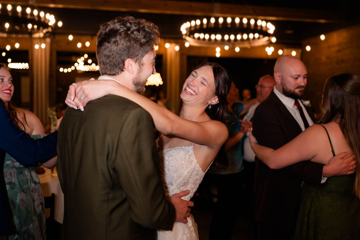 Bride in white gown laughing while dancing with groom in green suit at wedding reception.