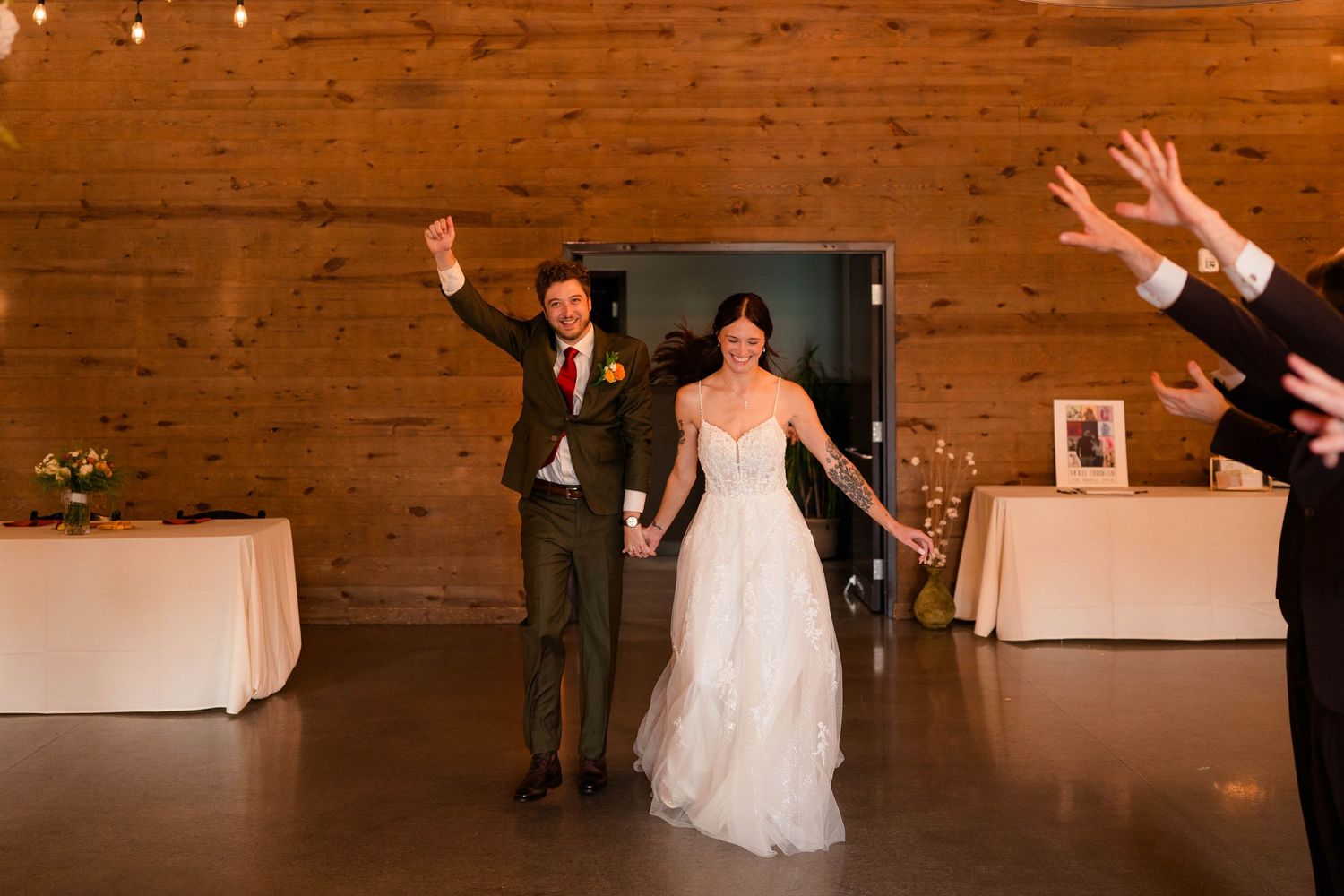 Bride in white lace gown and groom raise arms celebrating grand entrance into warmly lit rustic barn reception.