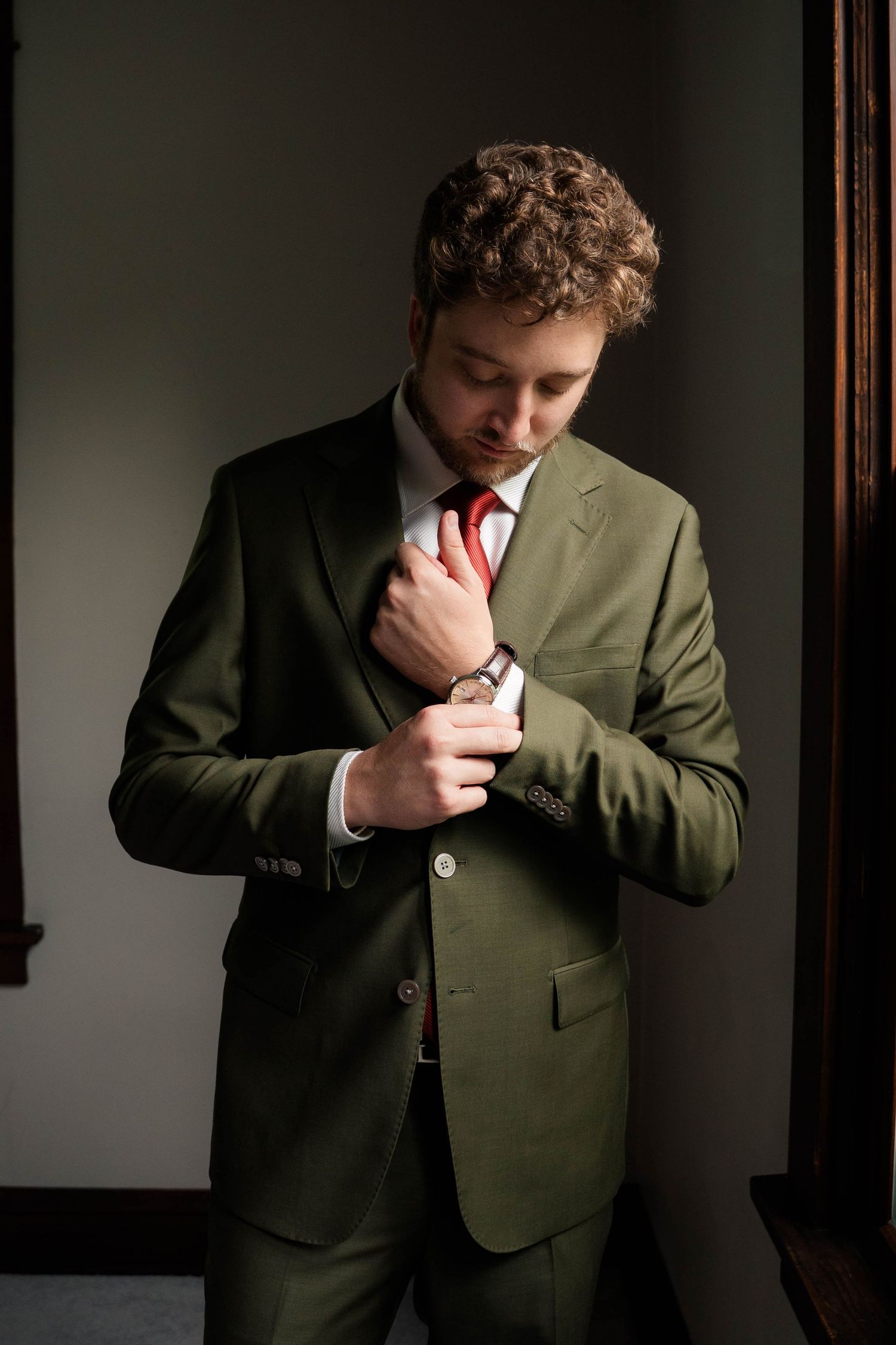 Stylish groom wearing olive green suit and red tie fastening his cufflinks before wedding.