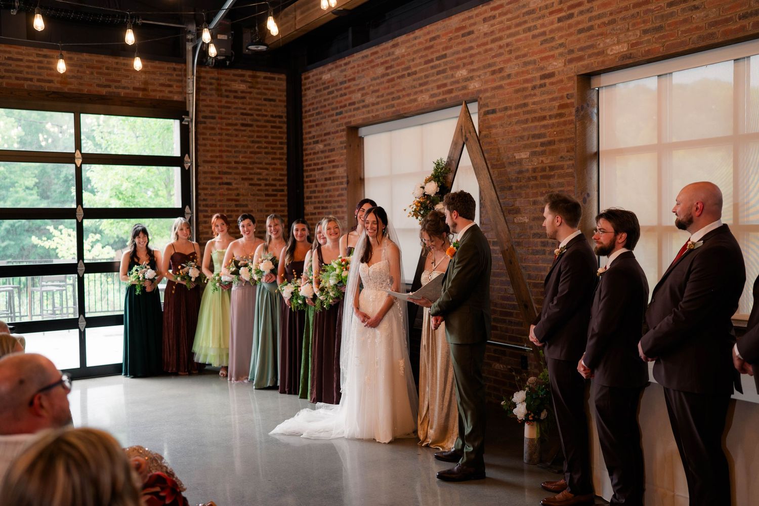 Wedding ceremony in brick venue with bride, groom, bridesmaids in green dresses, and groomsmen before triangular arch.