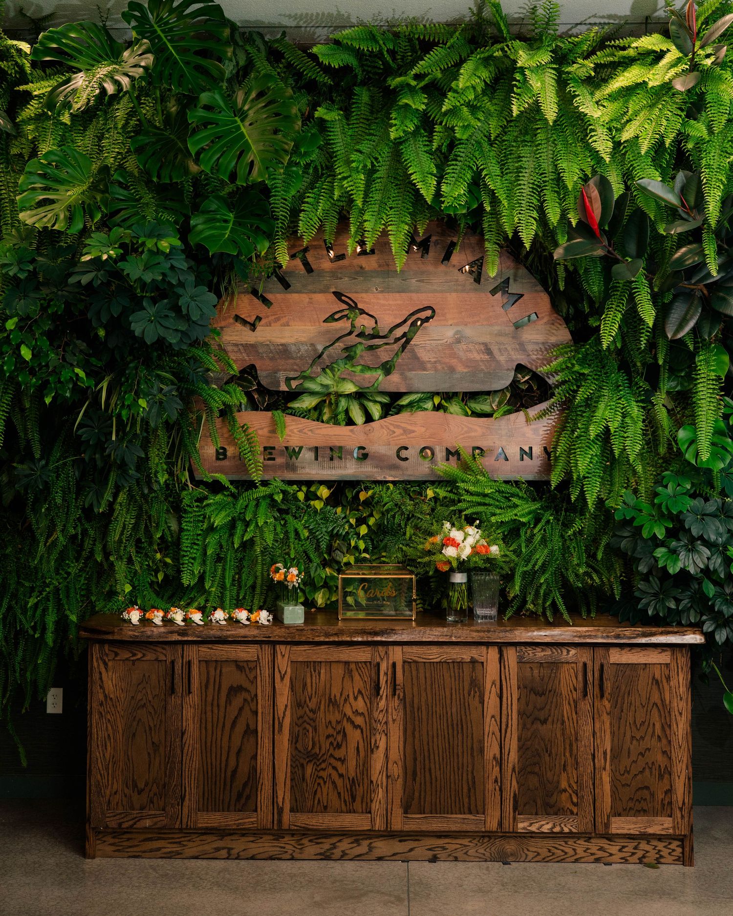 Lush green plant wall with wooden brewery sign and floral display on rustic wooden cabinet.