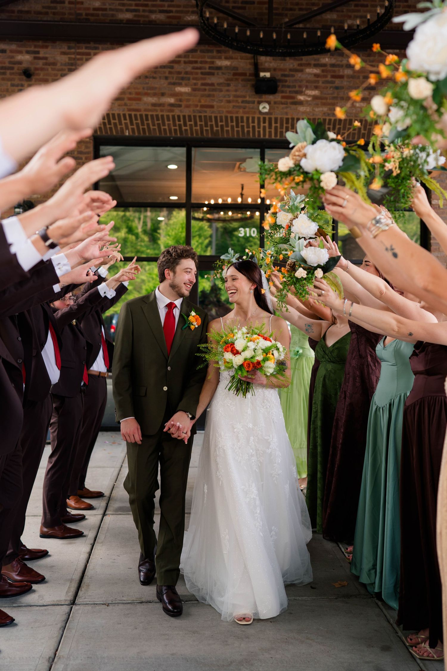 Bride and groom walk through a celebratory arch of raised hands and bouquets outside a brick wedding venue.