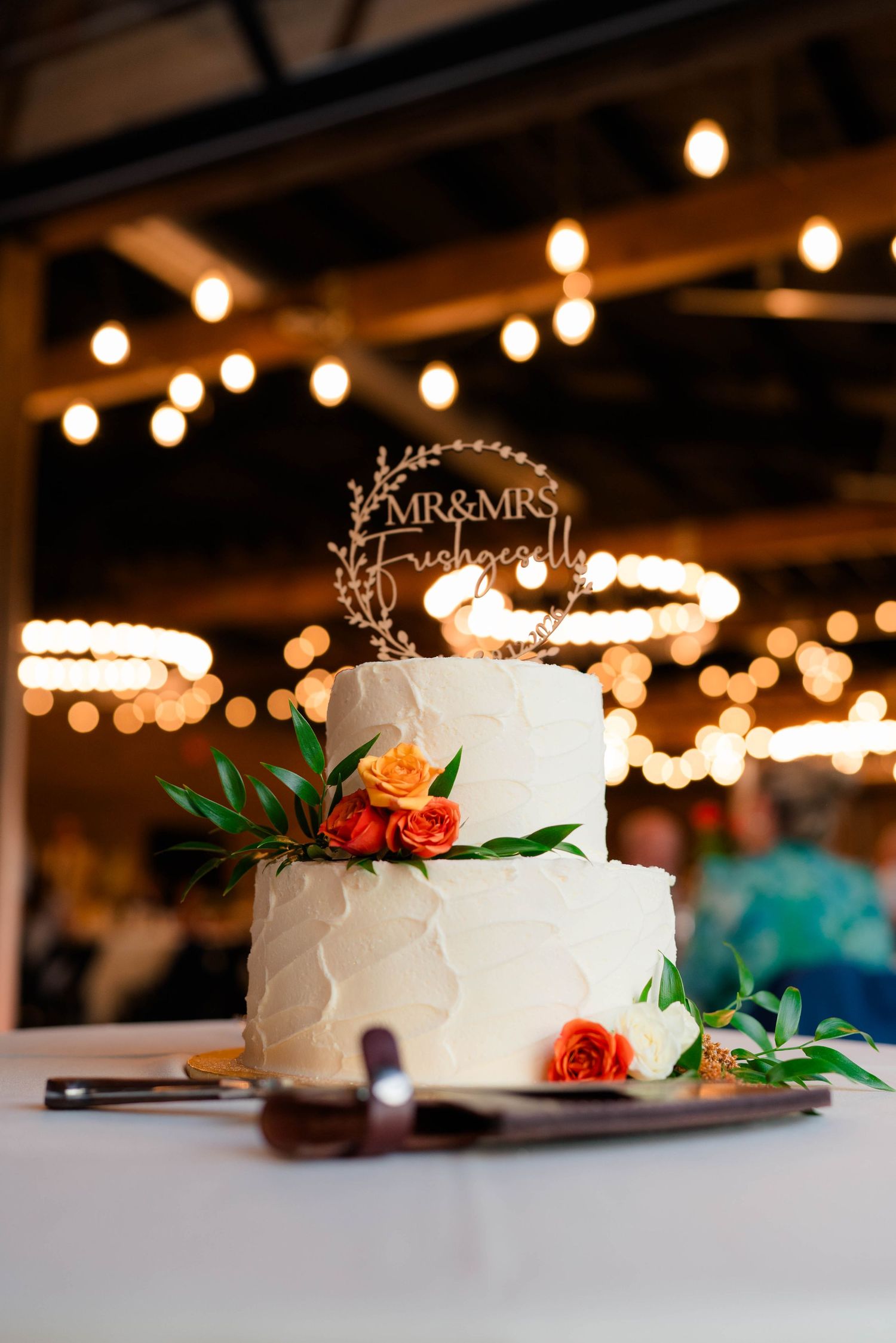 Two-tier white wedding cake with orange roses and Mr. and Mrs. topper under string lights.