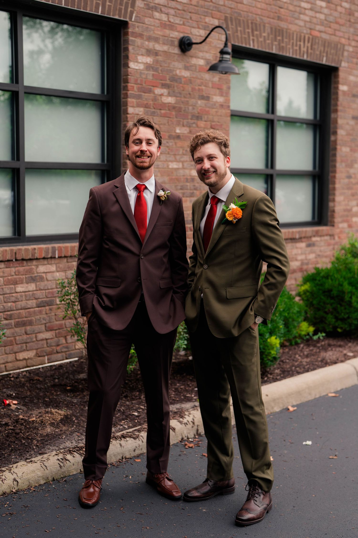 Groom in olive green suit and groomsman in dark brown suit smile together outside a brick venue.