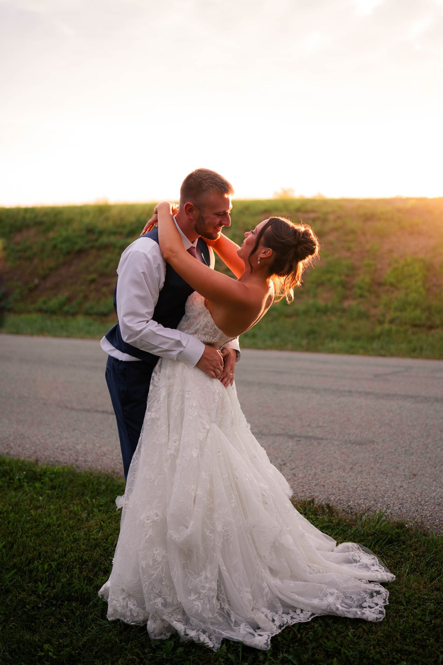 Bride and groom share a joyful embrace on a country road bathed in warm golden sunset light.